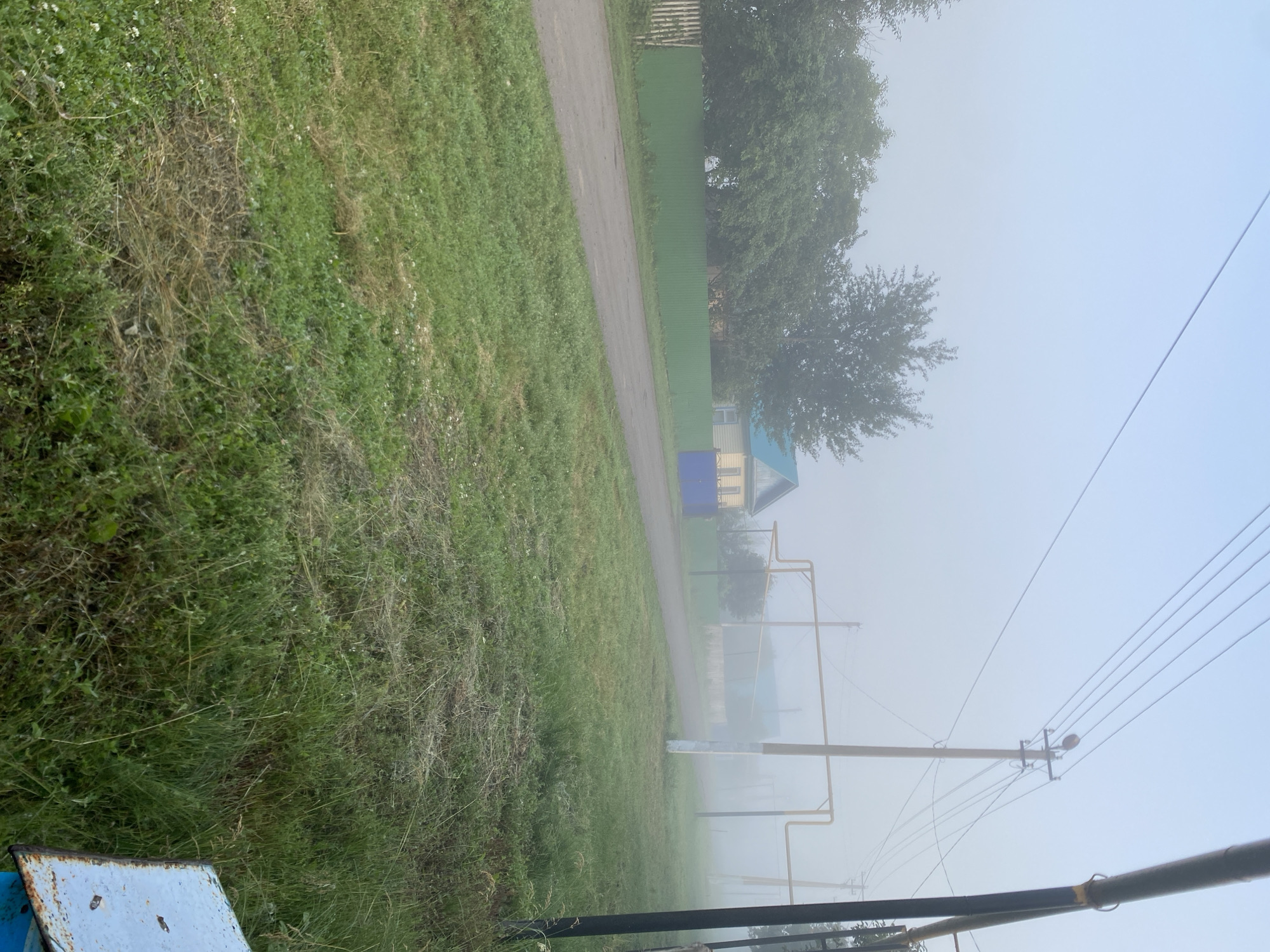 The image depicts a hazy rural scene in Maloarslangulovo, Russia, on a foggy morning. A dirt road runs through the foreground, bordered by green grass. In the middle ground, a green metal fence stretches across the frame, with a yellow house with a blue roof visible behind it. A blue gate is also present. Power lines crisscross the sky, supported by utility poles, and a yellow pipe runs overhead. The overall atmosphere is one of quiet and isolation, typical of a misty rural morning.