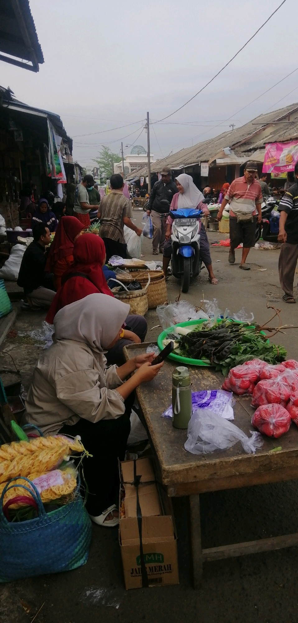 A street market scene unfolds in Ajibarang, Indonesia, under an overcast sky. People are engaged in various activities, with a focus on commerce and daily life. In the foreground, a woman wearing a light-colored hijab and jacket is seated at a wooden table, engrossed in her mobile phone. Beside her on the table are a green water bottle, plastic bags, a pile of tomatoes, and a tray of green vegetables including what appear to be long beans and paku leaves. To the left, a blue woven basket holds bundles of dried yellow noodles. Stacked cardboard boxes are visible to the lower left of the table.
Further into the scene, a blue and white motorcycle is parked, with a woman in a white hijab riding it, looking forward. To her right, a man in a striped orange and white shirt, wearing a fanny pack, walks away from the viewer. A man in a black jacket and cap is standing behind the woman on the motorcycle, and another man in a brown and white striped shirt is walking in the background carrying a plastic bag.
On the left side of the frame, several individuals are seated and standing near stalls with awnings. Some are wearing hijabs. The market appears to be set up along a narrow street lined with structures with corrugated tin roofs. The ground is paved. The overall atmosphere suggests a bustling local marketplace.
