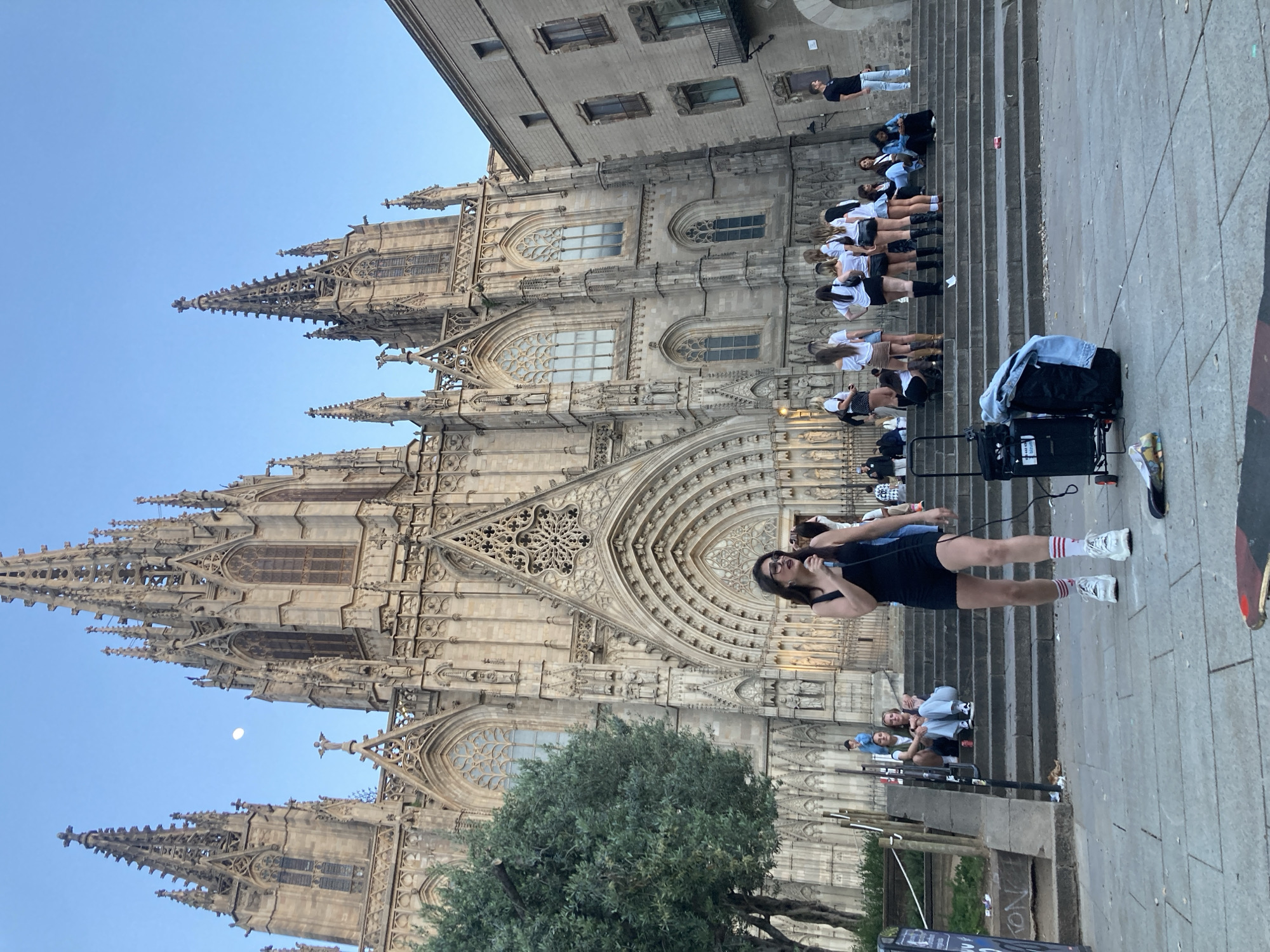 The image captures an outdoor scene in front of a grand Gothic cathedral, identifiable as the Barcelona Cathedral, distinguished by its intricate facade, spires, and prominent rose window. The image is rotated 90 degrees clockwise.
In the foreground, a young woman with dark hair, wearing a black top, shorts, white sneakers, and red-striped socks, stands holding a microphone. She appears to be speaking or singing to a group. Next to her on the ground are a black wheeled case, likely containing sound equipment, and a small speaker or amplifier.
Behind her, a group of approximately ten young women, many dressed in matching white t-shirts and dark shorts or skirts, are seated or standing on the wide stone steps leading up to the cathedral's main entrance. They face the woman with the microphone, suggesting they are an audience or participants in an activity. Further up the steps, and on an adjacent building to the right, additional individuals can be seen observing the scene.
The cathedral's detailed Gothic architecture, including numerous arched windows, flying buttresses, and decorative carvings, forms a majestic backdrop. A large green tree is situated in the mid-ground on the left, partially obscuring the lower portion of the cathedral. The sky is a clear, bright blue, with a distinct crescent moon visible, indicating either early morning or late afternoon. The lighting is soft and even. While there is some indistinct text on the black equipment case, it is not legible. The scene suggests a public performance, guided tour, or group gathering in a historic urban setting.