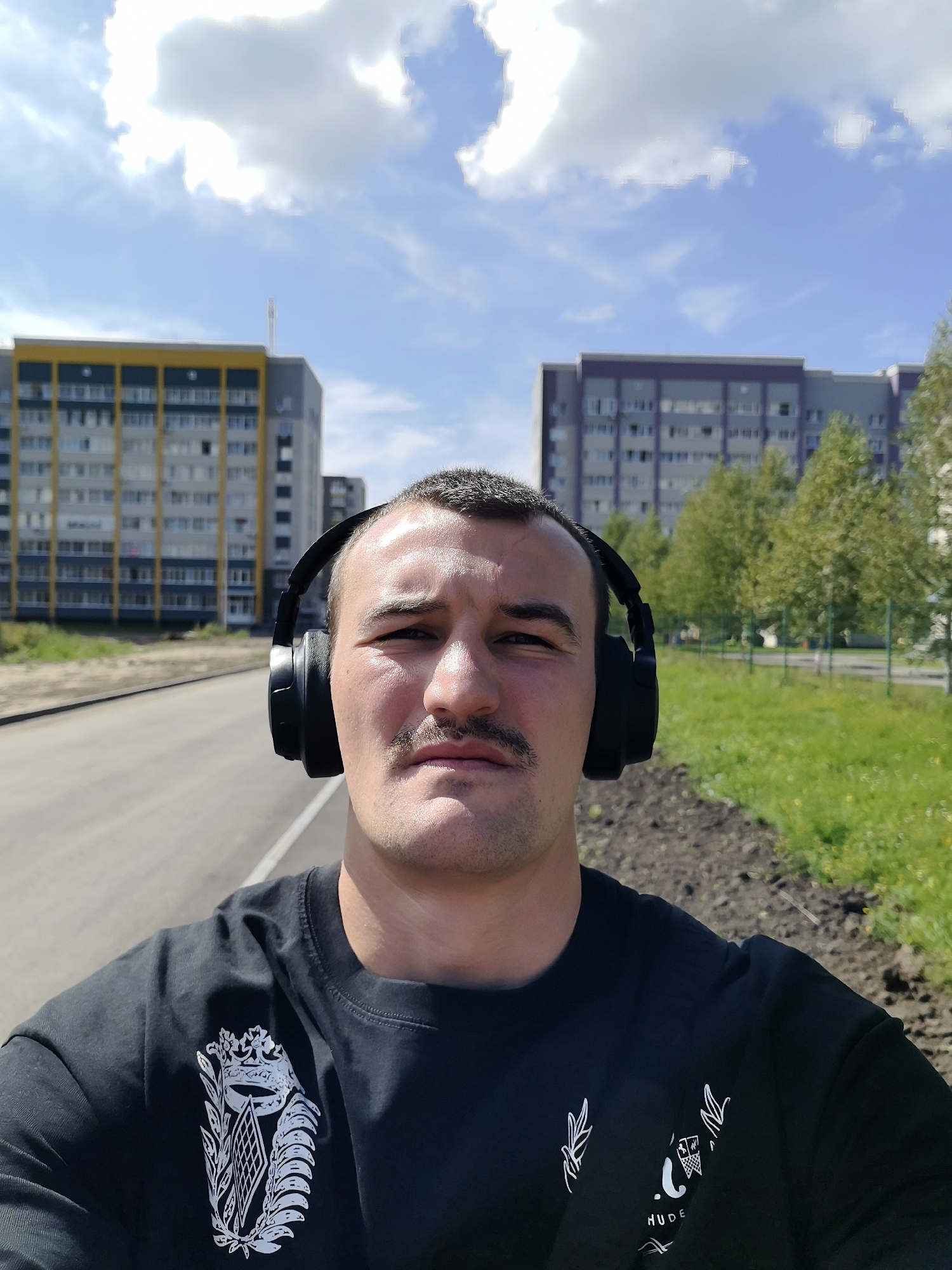 A medium shot selfie shows a man with a mustache wearing black headphones and a black t-shirt with a white emblem on the left chest. He stands outdoors on a paved surface in front of two apartment buildings, one yellow and gray, the other purple and gray. The sky is blue with white clouds. The ground to the right is covered with dirt and grass. The man is looking directly at the camera.