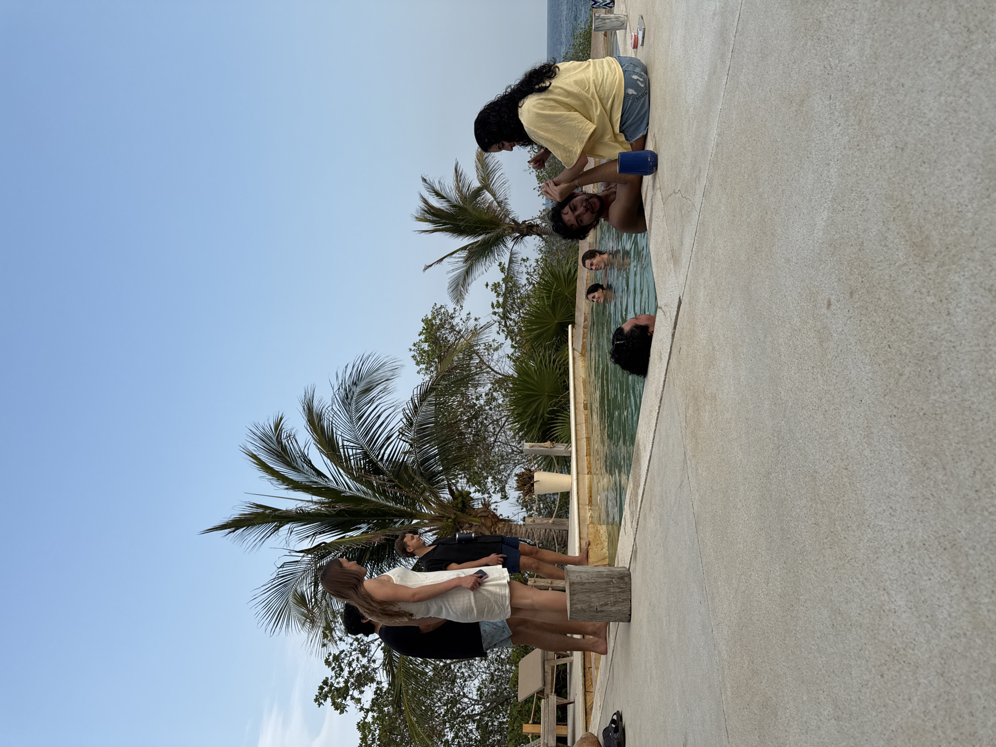 The image shows a group of people near a swimming pool outdoors. Some individuals are in the pool, while others are standing or kneeling at the pool's edge. One person is pouring something from a blue cup onto another person's head in a playful manner. Several people are standing near palm trees and other tropical vegetation. The sky is clear and blue. The location is Unknown, Mexico.
