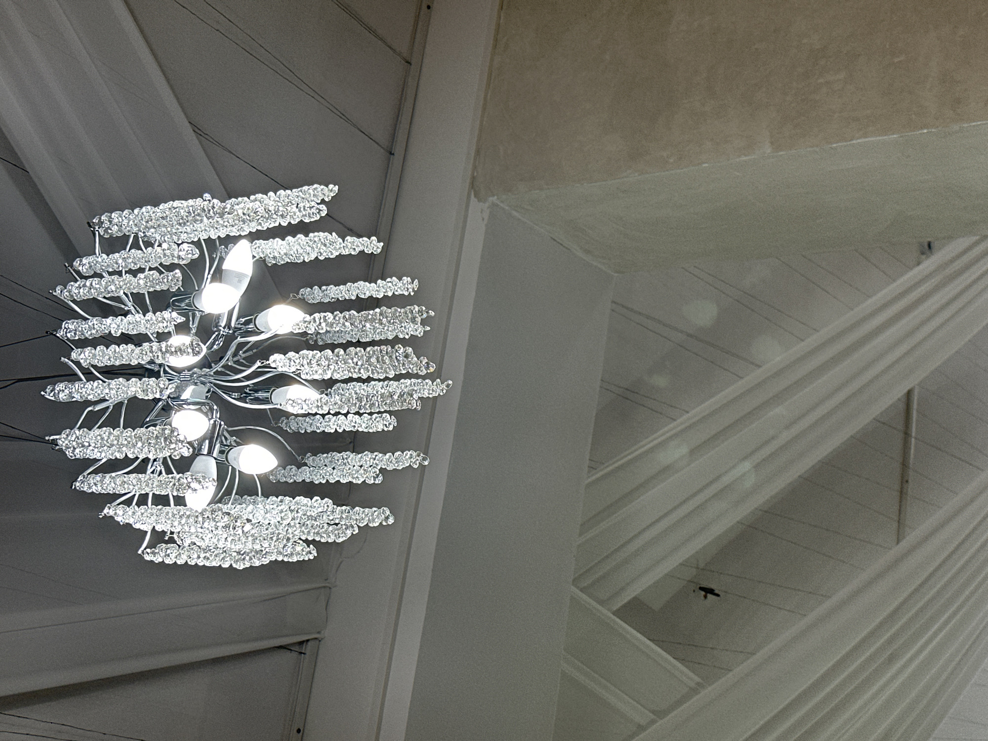 A low-angle shot captures a chandelier hanging from a ceiling. The chandelier has multiple tiers of crystal-like ornaments, resembling branches adorned with droplets, and several lights illuminate the surrounding space. The background features architectural elements with a combination of white draping fabrics, a smooth white column, and a textured gray ceiling section. The image focuses on the juxtaposition of the ornate chandelier with the modern or abstract ceiling design.