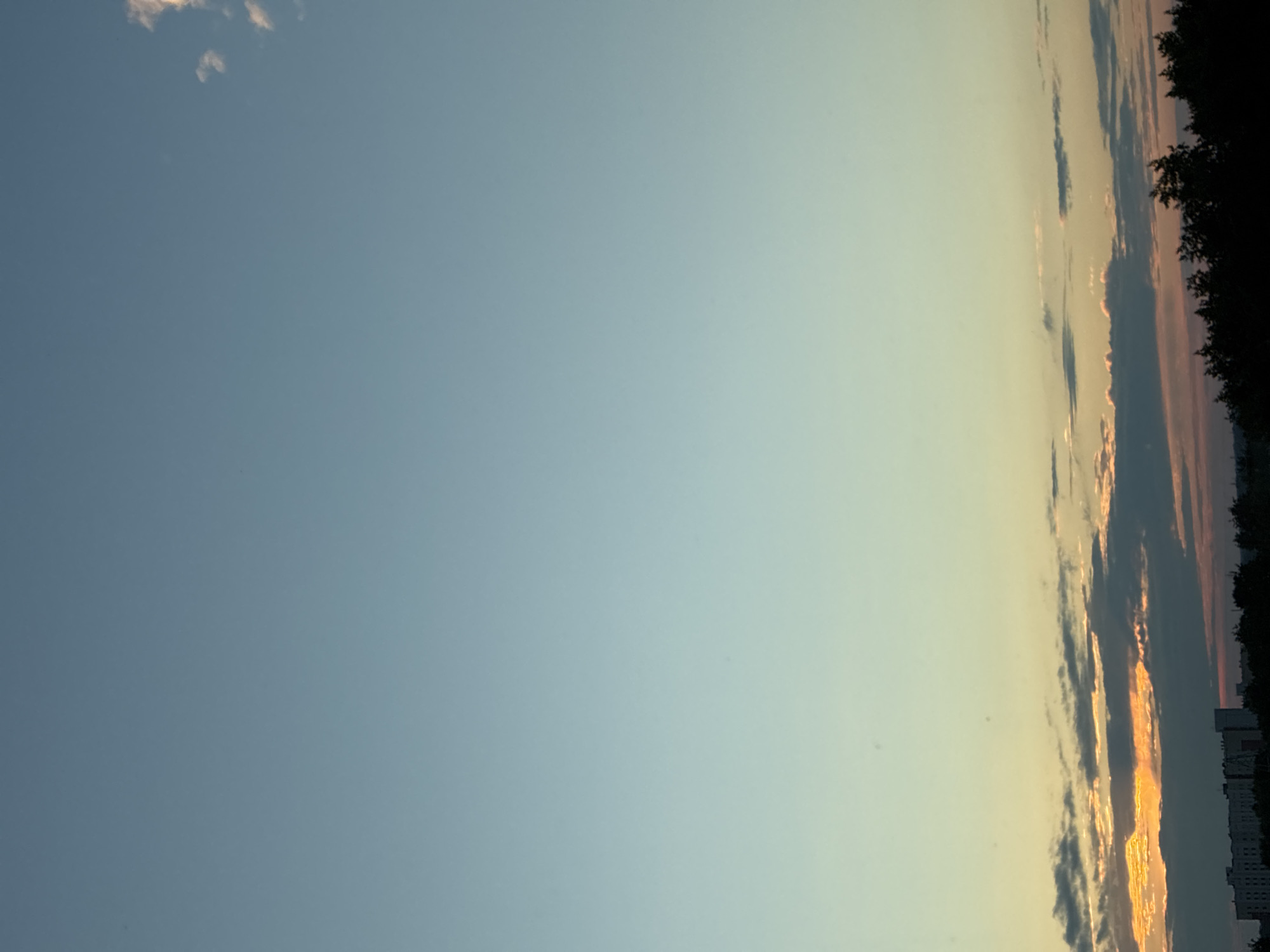 A vertical, full-frame, outdoor photograph captures a serene sky at dusk over the city of Unknown, Unknown. The sky transitions from a pale blue at the top to a softer, yellowish hue near the horizon. A few wispy clouds are scattered across the upper portion of the sky, with more prominent, darker clouds lining the horizon. Beneath the clouds, a sliver of warm orange light indicates the setting sun. The lower edge of the frame shows a silhouette of trees and buildings, suggesting an urban landscape. No people or specific actions are visible.