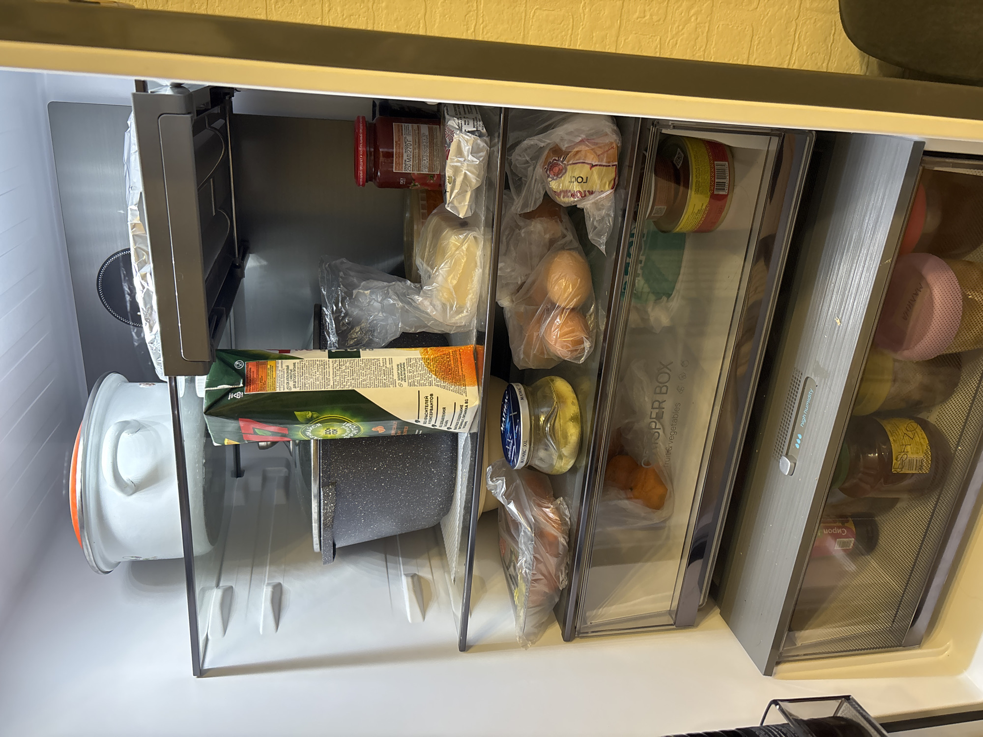 Here is a matter-of-fact description of the image:
The image shows the interior of a refrigerator. The refrigerator is stocked with various food items. Visible items include a covered cooking pot, a carton of juice, a jar of tomato sauce, cheese, a loaf of bread, eggs, pickles, carrots, and several jars of preserves or other canned goods. The refrigerator has a crisper drawer labelled "Crisper Box" for fruits and vegetables. The labels are in Cyrillic, suggesting a location in Russia, possibly Верхнеказымский. The inside of the fridge is lit with a cool light.