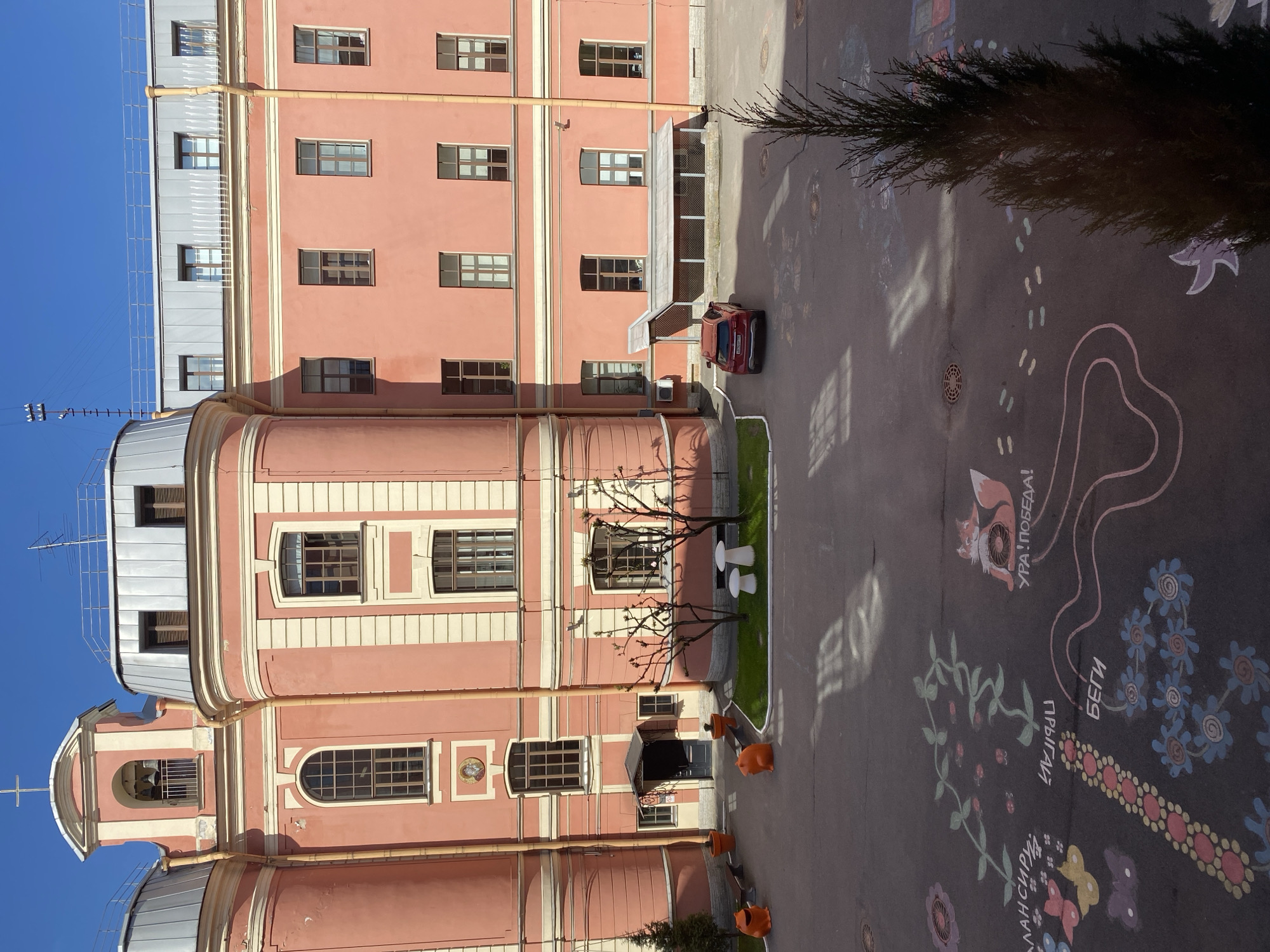 The image shows the courtyard of the Smolny Cathedral in Saint Petersburg, Russia. The courtyard is paved with asphalt and features chalk drawings on the ground. A red car is parked near a building entrance. Various objects are in the scene, including orange traffic barriers, outdoor tables, and landscaping. The architecture is distinctive, with a light-pink building displaying multiple windows and ornamentation. The sky is blue.