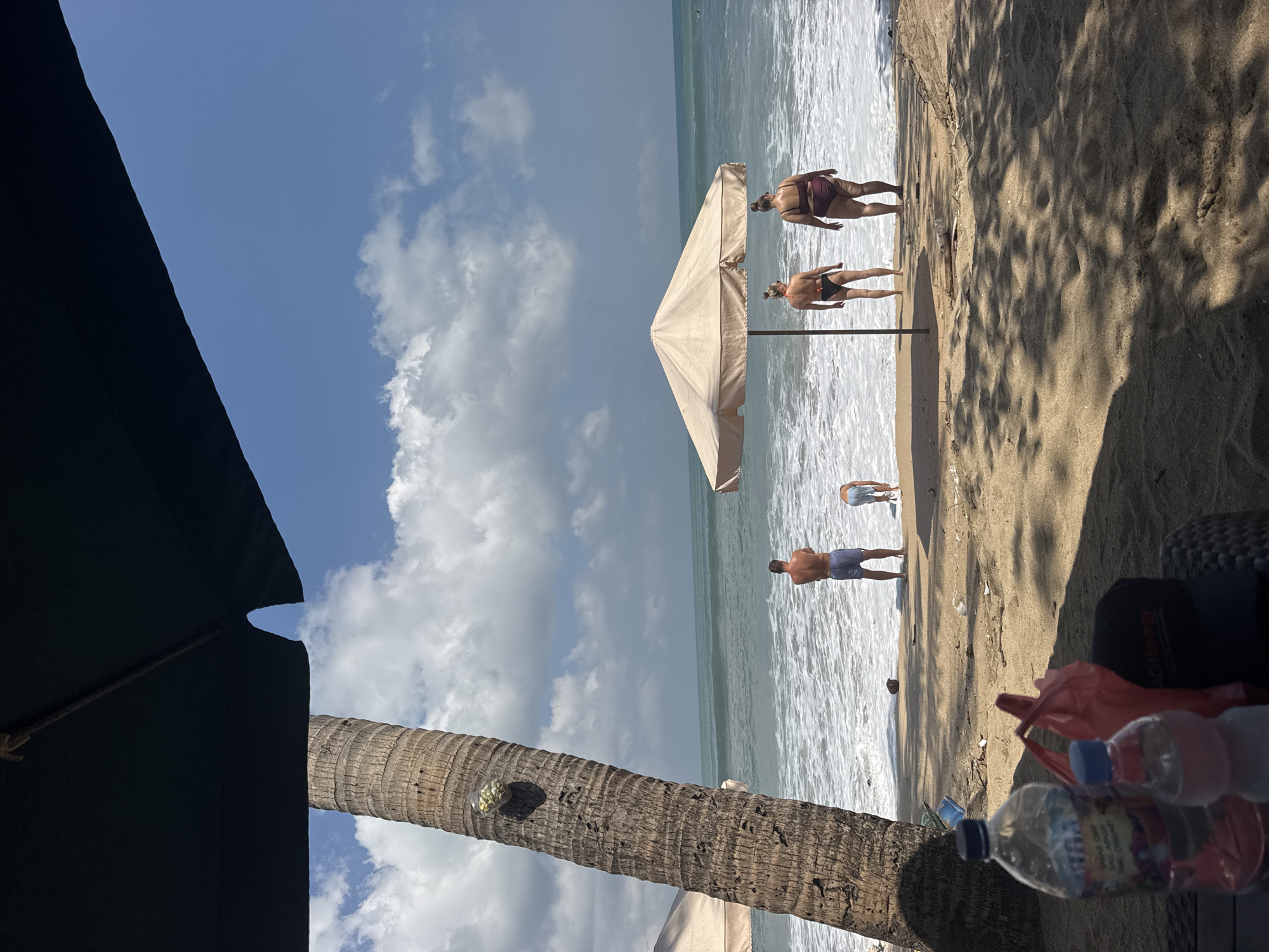The image depicts a sunny day on Kuta Beach, Indonesia. In the foreground, two plastic water bottles and a red bag are placed on a table, casting shadows on the sand. A large palm tree trunk is visible on the left, partially covered by a dark umbrella. In the mid-ground, the sandy beach meets the ocean with gentle waves. Three adults and one child are present. A man in blue shorts stands facing the ocean. A child in light-colored shorts is bent over near the water's edge. Two women in bikinis stand facing the ocean, one in a black bikini and the other in a burgundy bikini. A light-colored beach umbrella is set up near the women. The sky is blue with scattered white clouds. The ocean is a turquoise green.