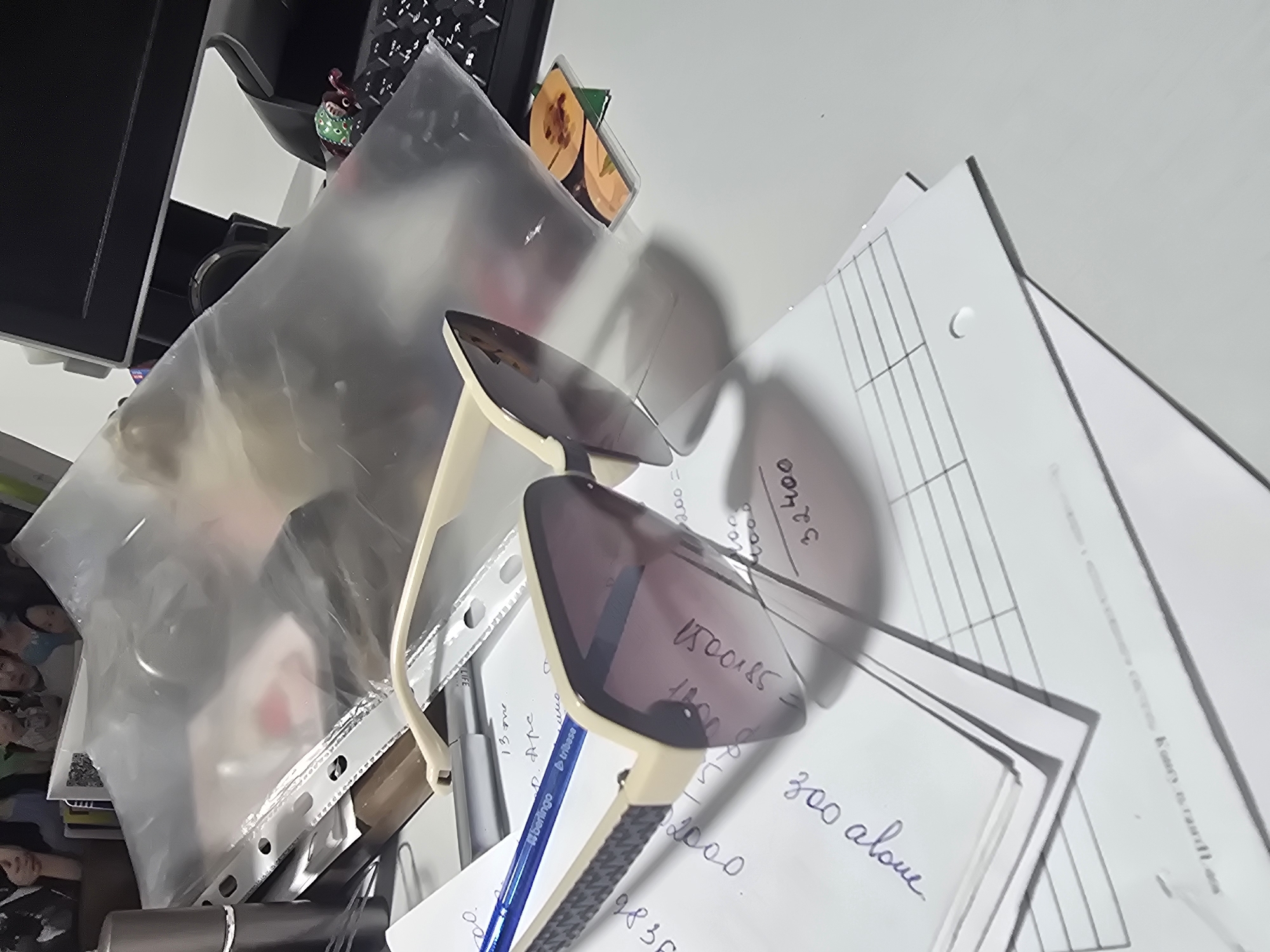 A close-up shot focuses on a desk cluttered with various items, suggesting a workspace. In the foreground, a pair of cream-colored sunglasses with dark lenses rests on a stack of papers. One lens reflects a portion of the environment, showing some handwriting. A blue pen with "berlingo" and "tribase" branding lies on top of the sunglasses.
Behind the sunglasses, a clear plastic folder filled with what appears to be photographs or documents is propped up, obscuring the background. The folder contains a few loose items, including what looks like a small, decorative object with green and brown accents on the right side.
To the left, a silver cylindrical object, possibly a perfume atomizer or a flashlight, stands upright. Various papers with handwritten notes and printed grids are scattered across the desk. Some of the handwritten notes are partially visible and include numbers like "32400" and "300 alone".
The desk surface appears to be a light gray laminate. The overall lighting is somewhat dim, with shadows cast by the objects. The scene is likely indoors, possibly an office or a home desk. The specific location in Ulan-Ude, Russia, cannot be determined from the visual information provided in the image.