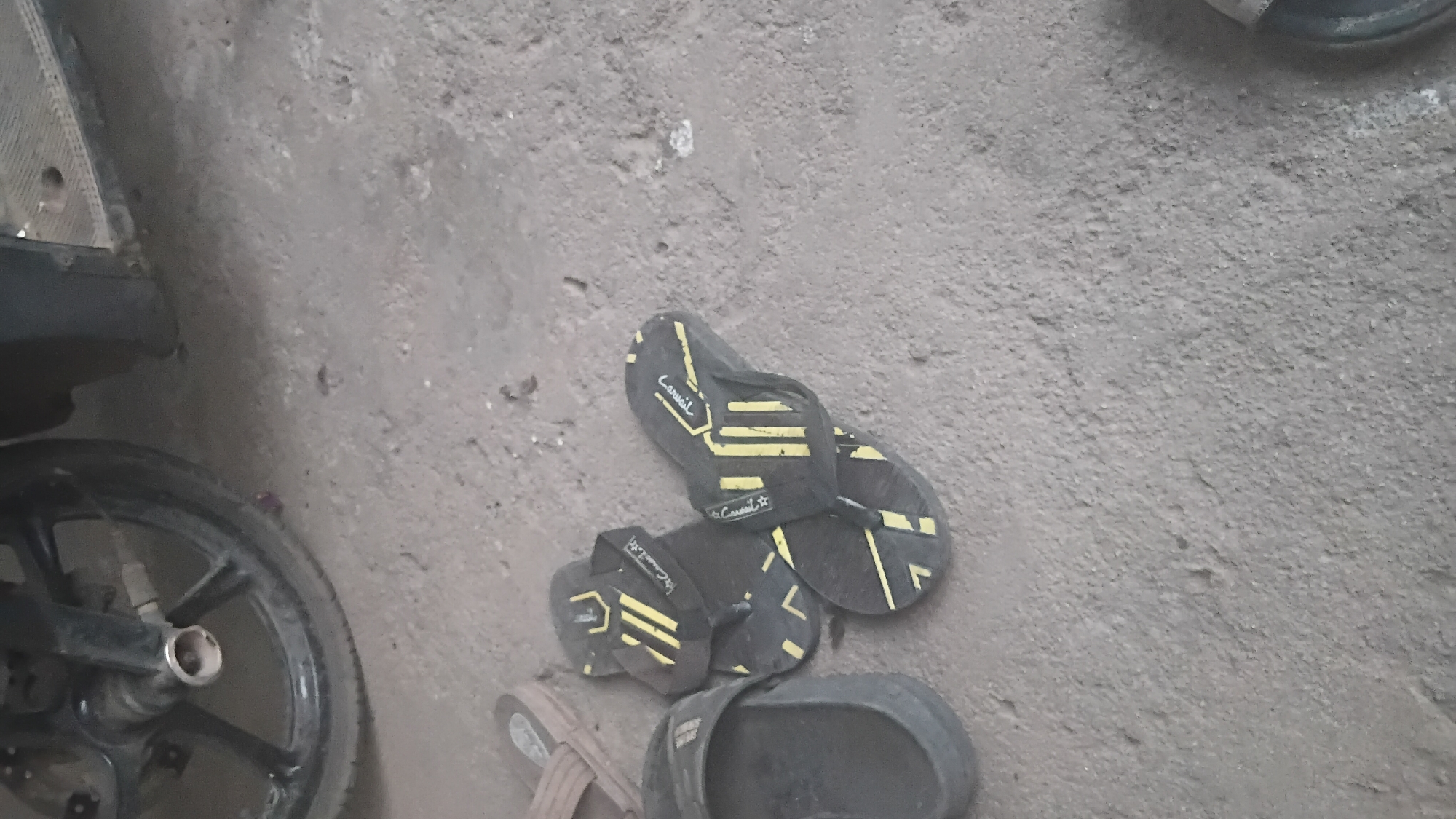 This is a low-angle, close-up shot of several pairs of sandals and flip-flops scattered on a dusty, concrete ground, with the wheel and part of a motorcycle visible in the upper left corner. The lighting is dim, suggesting an indoor or shaded outdoor environment. The scene appears to be in Prabumulih, Indonesia.
The foreground is dominated by the footwear. To the left, a dark grey, slip-on sandal is partially visible. In the center, two pairs of black flip-flops with yellow striped accents are laid out. The flip-flops have a distinctive design with diagonal yellow stripes on the soles and on the straps. The brand name "Casual" is visible on the straps of these flip-flops. Further back and to the left, a light brown sandal is also present.
The ground is composed of rough, granular concrete or compacted dirt, with scattered small stones and debris. The motorcycle wheel is black with silver spokes and brake components. The overall impression is one of everyday, utilitarian objects left in a common area, possibly a workshop, garage, or outdoor patio.