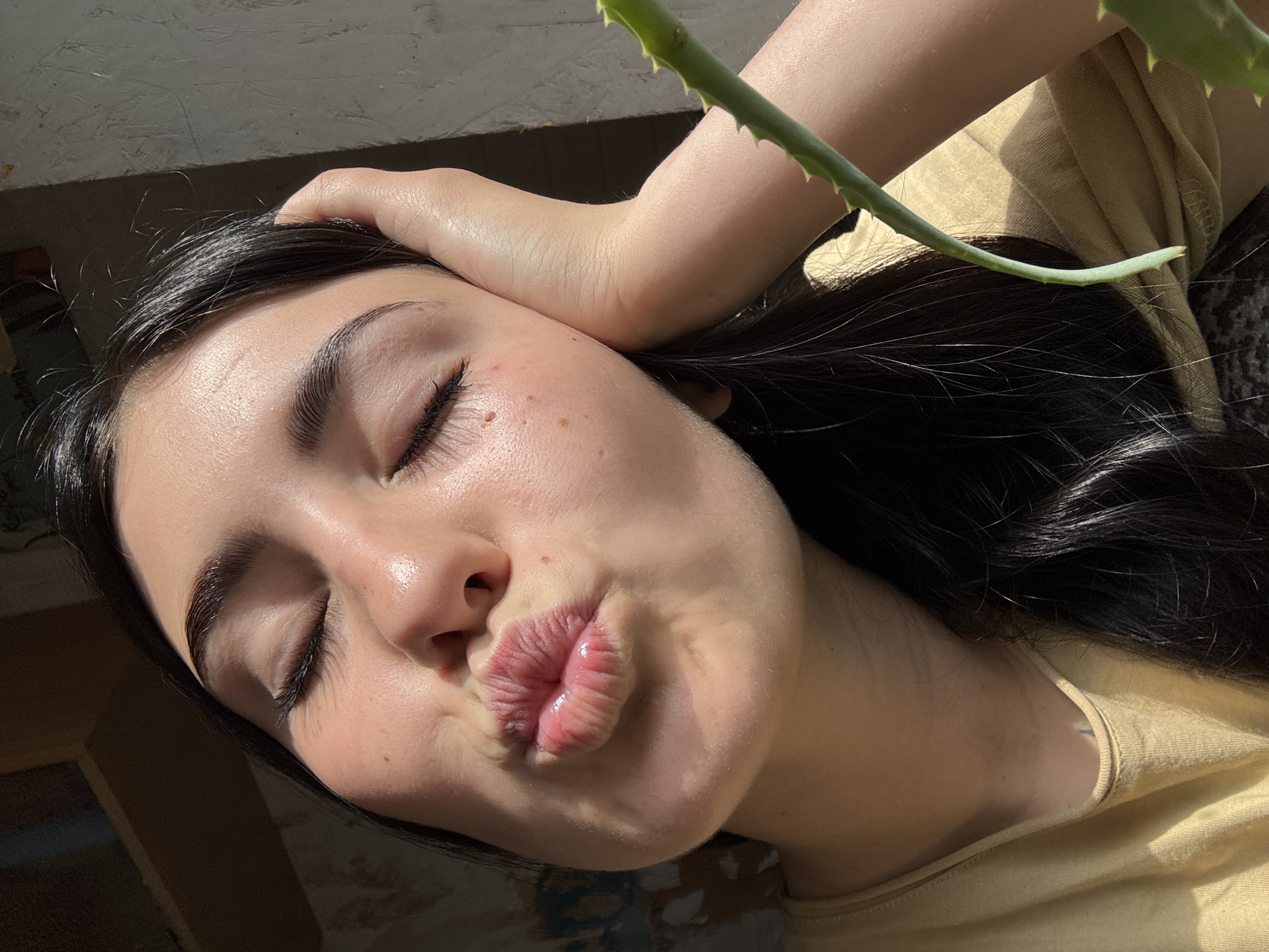 In the city of Fokino, Russia, a young woman with dark hair is captured in a close-up, sunlit selfie. Her eyes are closed, and her lips are pursed in a kissing gesture. Her right hand is raised, supporting her head, with her fingers resting on her temple. A green succulent plant stem is visible in the foreground, to the right of her face. The background is dimly lit, with hints of shelving or furniture on the left and a textured wall on the right, suggesting an indoor environment with natural light streaming in from a window. The woman is wearing a light yellow t-shirt.
