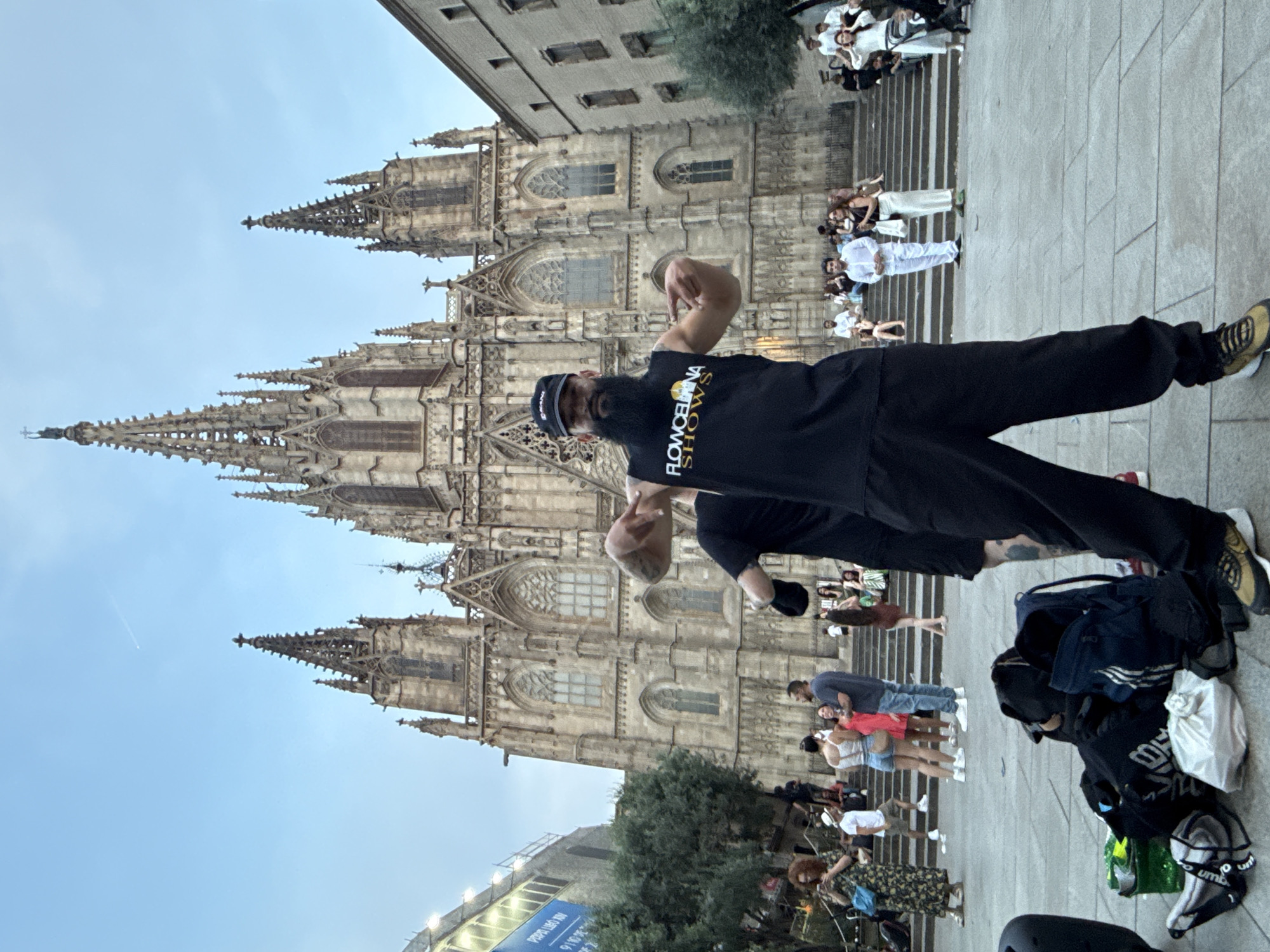 A man stands in front of the Barcelona Cathedral in Spain, posing with his arms raised to display his biceps. He wears a black t-shirt with yellow lettering that reads "FLOWCELONA SHOWS," black pants, and gold sneakers. To his right, a pile of bags and a helmet sit on the ground. In the background, people are sitting on steps leading up to the cathedral and also walking nearby. The sky is pale blue, and the cathedral is a large stone structure with intricate details. A street sign is visible at the bottom of the image.