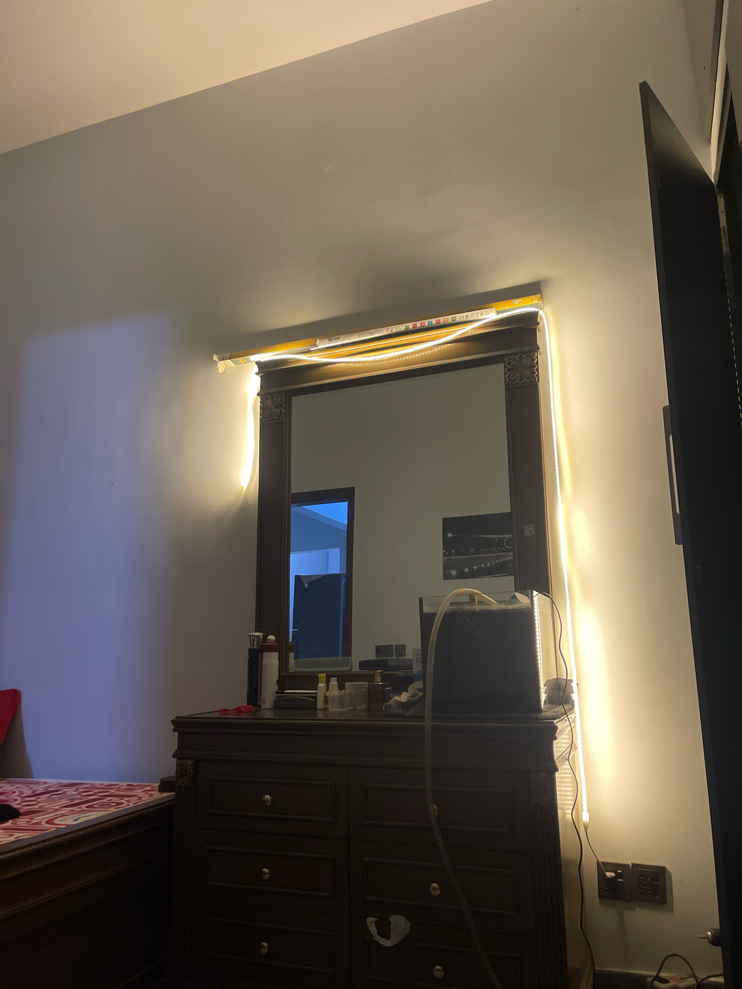 The image depicts a bedroom interior. A large, dark wood dresser with a mirror is centrally placed against a white wall. LED strip lights are affixed to the top and sides of the mirror frame, casting a warm glow on the wall. The mirror reflects a doorway leading to another room, with a blue interior. On top of the dresser are various items, including bottles, a black object that appears to be a computer monitor, and some tubing. To the left of the dresser, part of a bed with a red and white patterned comforter is visible. The overall impression is of a bedroom that is illuminated by decorative lighting. The image does not provide any specific indicators of its location within Karachi Division, Pakistan.