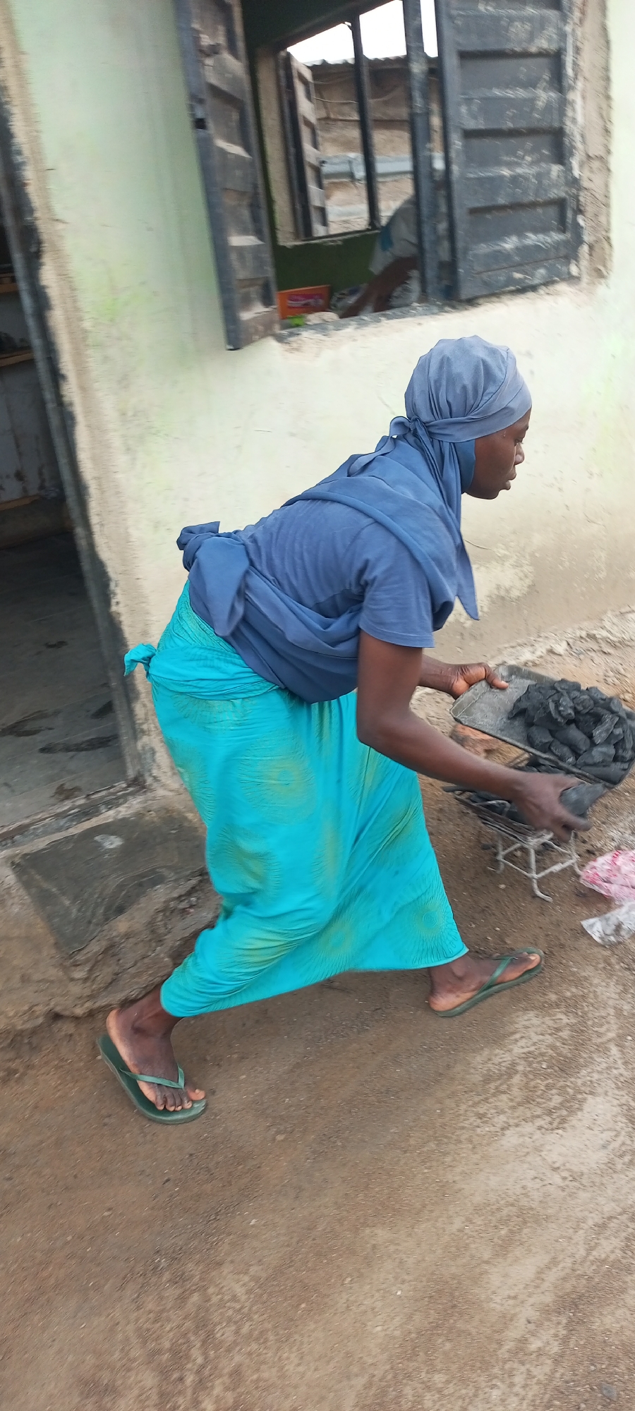 A woman stands on an earthen surface in Adankolo, Nigeria. She wears a headscarf, a blue shirt, and a long turquoise skirt. She's bent over and holding a metal tray filled with charcoal. She is using her hand to place the charcoal into a grill. Behind her is a building with a window and an open doorway. Another person can be seen through the window.