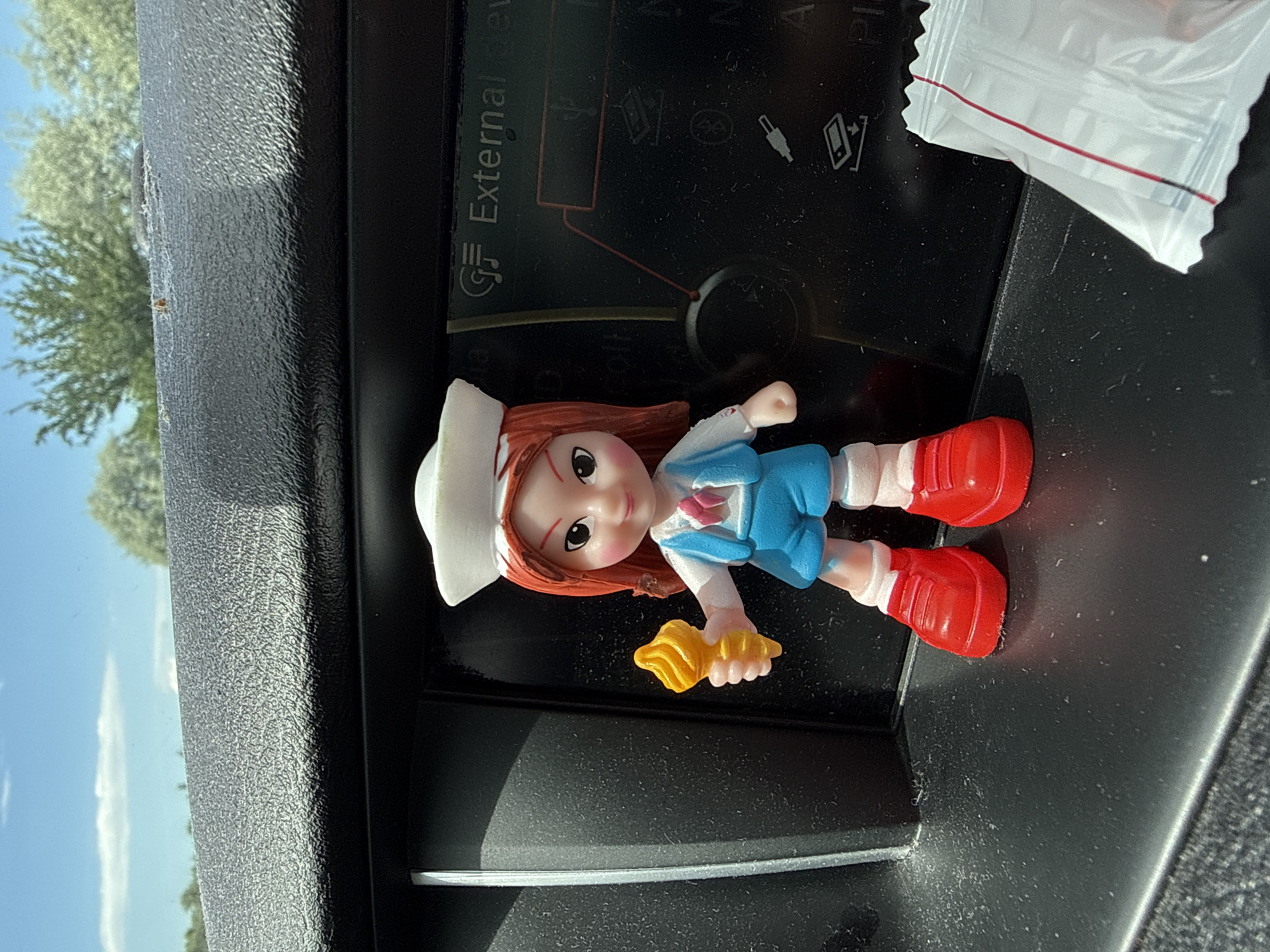 A brightly lit, slightly tilted photograph captures a small figurine placed on a textured, dark surface within a vehicle's interior. The figurine depicts a girl with reddish-brown hair, a white cap, a light blue top with white sleeves, and red boots. She has a gentle smile and is holding a yellow, shell-shaped object in her left hand.
The figurine rests on what appears to be a car's dashboard or center console. Behind it, a dark panel or screen displays various icons, including symbols for USB and Bluetooth, and the partially visible word "External" in white text. To the right of the figurine, a corner of a small white plastic packet with red and black striped edges is visible.
Through the car window on the left, a clear blue sky dominates the outdoor background, featuring a thin, elongated white cloud. Green foliage from trees is visible, suggesting an outdoor setting. The strong, direct light source indicates it is daytime with sunny weather. The scene is static, showcasing the figurine in this particular setting, located in Radinovo, Bulgaria.