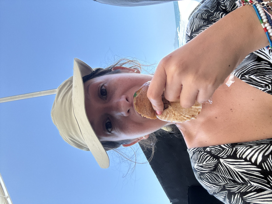 An outdoor, eye-level image captures a female individual, approximately middle-aged, aboard a vessel under a clear blue sky, identified as being in the city of Pak Nam, Thailand. The individual is centered in the frame, wearing a light-colored sun hat and a black and white patterned V-neck top with a leaf motif. She is actively consuming a round, brown-crusted food item, holding it in her right hand with a partial bite taken. The food is cradled in a fluted light-brown paper wrapper, partially peeled down. Green contents are visible within the bite mark. The individual wears small, circular stud earrings and multiple colorful beaded and metal bracelets on her left wrist. In the background, the sky is uniformly blue and clear. To the right, open water with visible white-capped waves indicates a body of water such as an ocean or sea, with a faint, distant landmass on the horizon. Elements of the vessel's structure, including a dark panel on the left and a white support pole on the upper right, are visible.