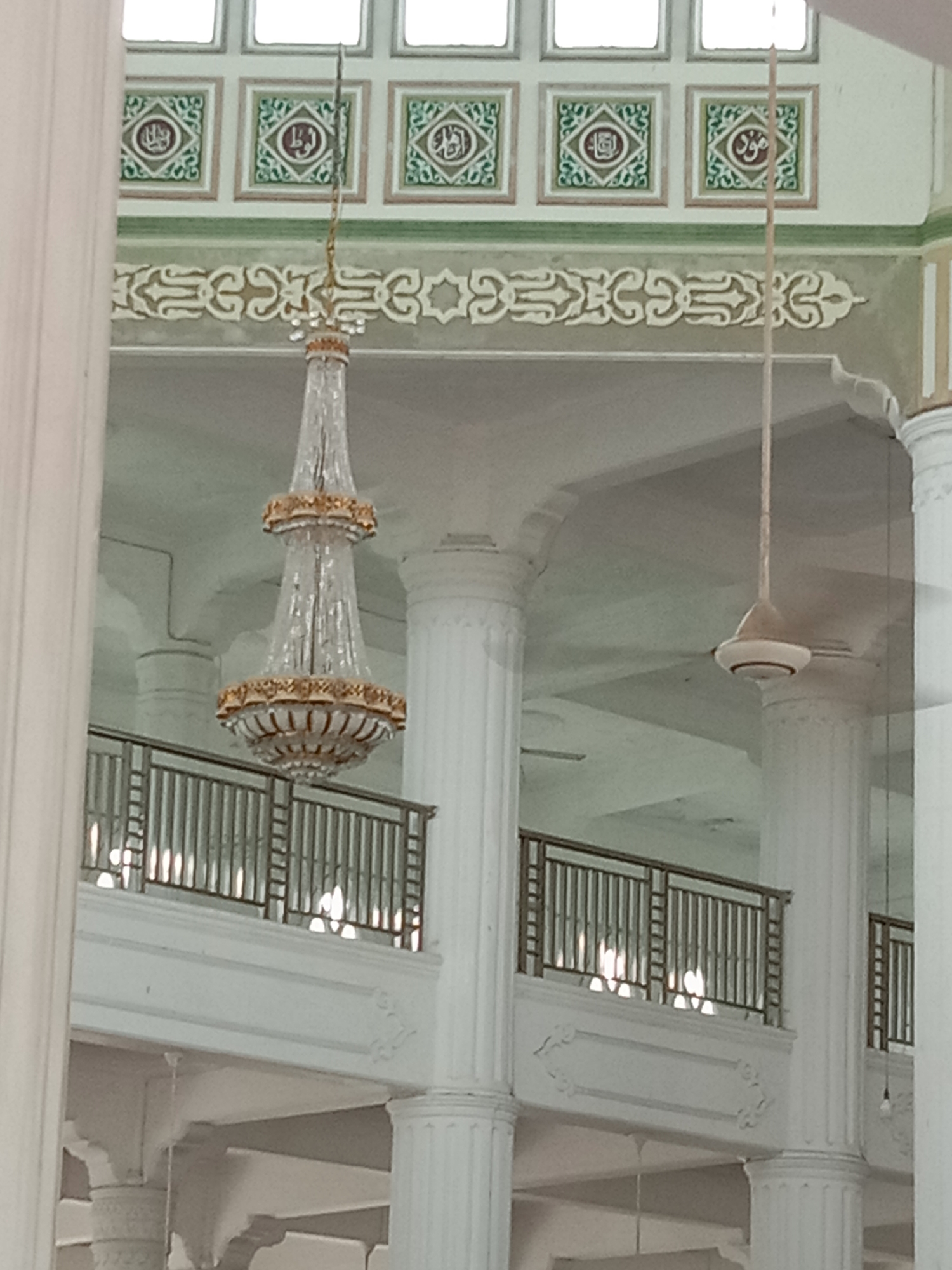 The image shows the interior of a grand building with high ceilings, characterized by ornate architectural details and decorative elements. In the foreground, a large, crystal chandelier with gold accents hangs prominently, suggesting a formal or religious setting. To the right, a ceiling fan is visible, indicating the presence of climate control systems.
The walls are adorned with decorative Islamic calligraphy within square frames, bordered by green and beige accents. Below this, a section of the wall features intricate, cream-colored scrollwork. The building has multiple levels, with balconies visible on the upper floors, complete with metal railings. Large, fluted white columns support the structure, adding to the sense of grandeur.
The lighting in the image appears to be a combination of natural light filtering in from above, likely from windows or skylights, and artificial lighting from unseen sources that create reflections on the balcony railings. The overall impression is one of elegance and a well-maintained, spacious interior.
While the image depicts an interior, the architectural style, particularly the Islamic calligraphy and the overall grandeur, strongly suggests a mosque or a similar religious institution. Given that the request specifies the city of Lhokseumawe, Indonesia, this building is likely the Islamic Center of Lhokseumawe (Masjid Agung Islamic Center Lhokseumawe). This landmark is known for its impressive architecture and serves as a central place of worship and community activity in the city.