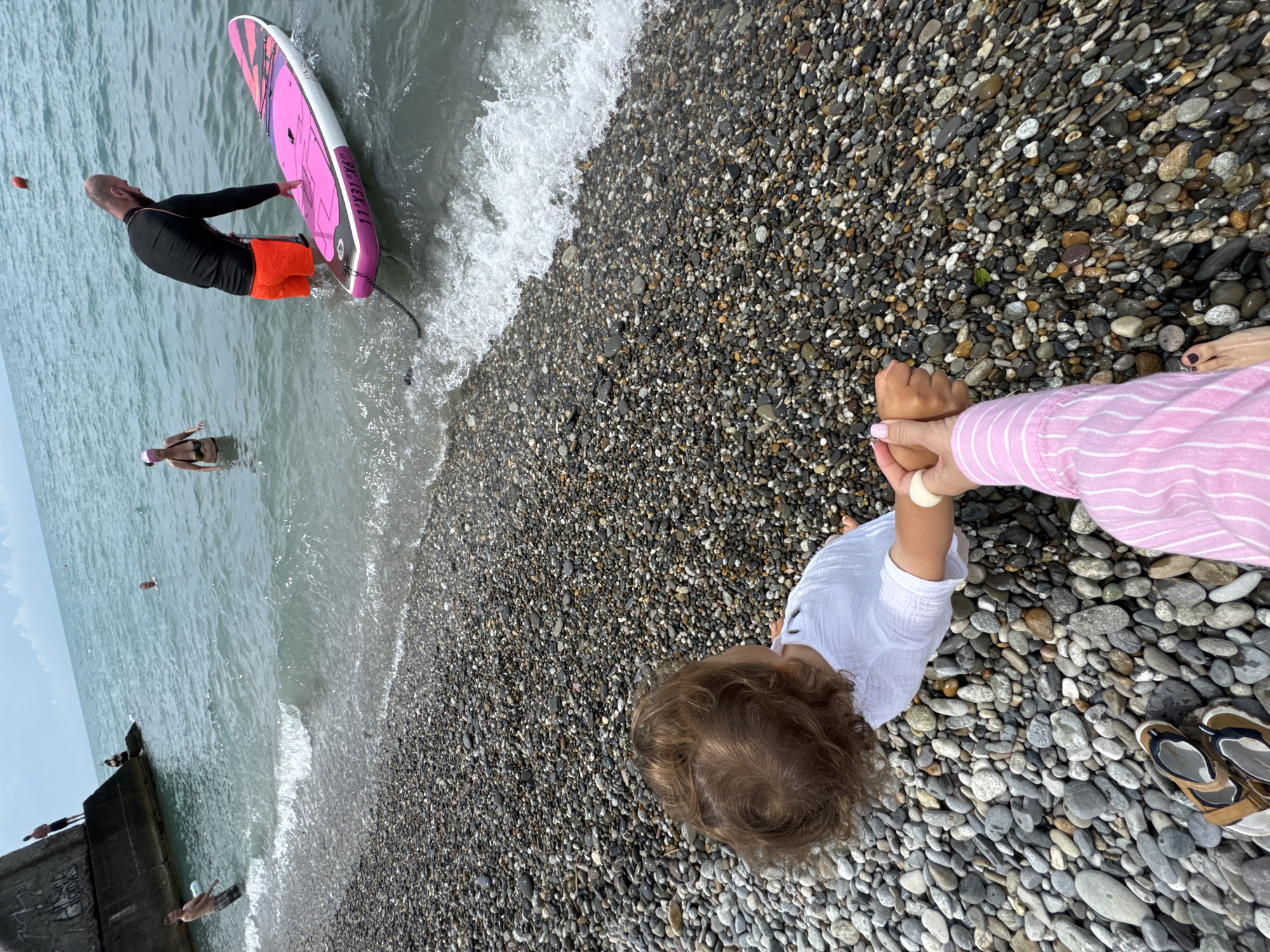 In Sochi, Russia, on a pebbled beach, a child with curly brown hair sits on the shore, wearing a white shirt. An adult, seen from the torso down, is seated next to the child, wearing a pink and white striped shirt. The adult's left hand holds the child's right hand. Several people are in the water in the background. One person is standing near the shore, holding a pink and black stand-up paddleboard. Another person is swimming in the distance. A concrete structure juts out into the sea on the left side of the image. The sky is overcast, and the sea is a muted blue-green.