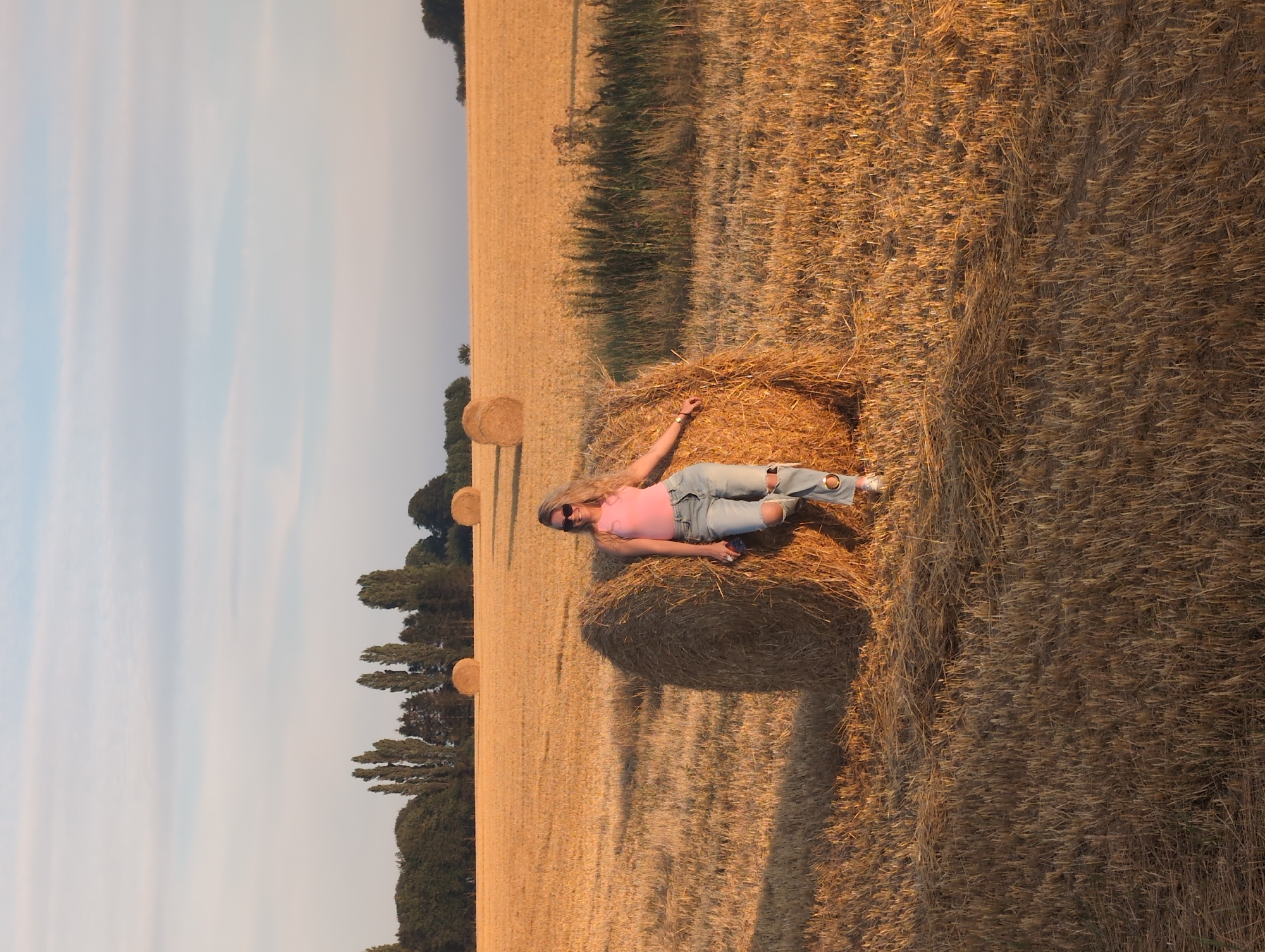 In Alvimare, France, a woman with blonde hair and sunglasses stands in a field of harvested wheat during sunset. She is leaning against a large bale of hay, wearing a pink tank top and ripped blue jeans. The field stretches out behind her, dotted with several more hay bales and a line of trees in the distance. The sky is a soft blue with wispy clouds, and the setting sun casts a warm, golden light over the landscape.