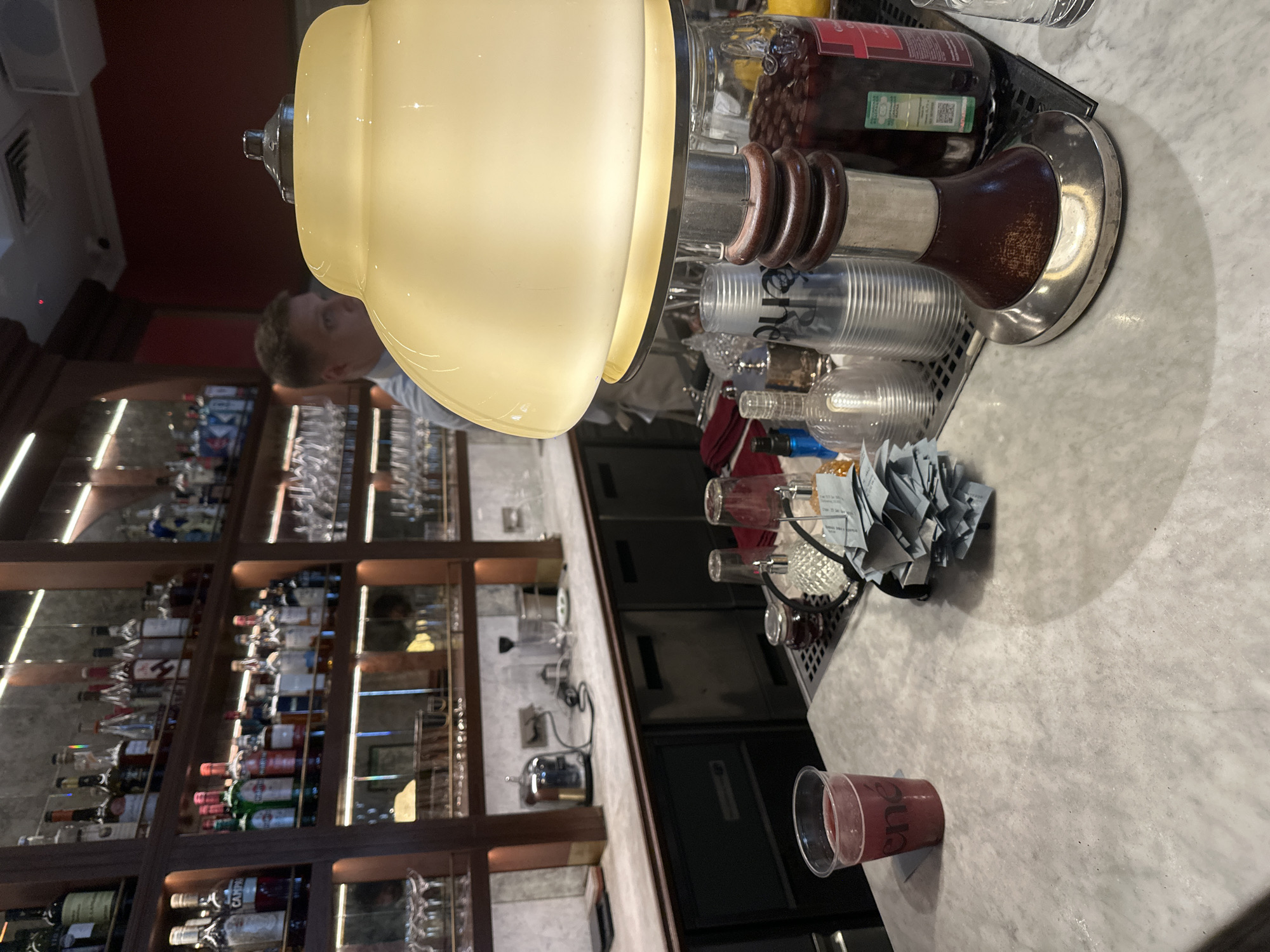 The image depicts a bar counter in a dimly lit establishment. In the foreground, a marble countertop occupies the lower portion of the frame. On the left side of the counter, a clear plastic cup containing a reddish beverage with the embossed letters "ene" is visible. To the right of the cup, a stack of light blue slips of paper, possibly receipts or tickets, is piled. Behind this, a metallic ribbed cup with the word "ene" printed on it stands next to a glass jar filled with red cherries. A vintage-style lamp with a cream-colored shade and a dark wooden base is positioned prominently in the center foreground, casting a warm glow. In the background, shelves are stocked with various bottles of alcohol, and a man in a light-colored shirt is partially visible, seemingly attending to the bar. The overall ambiance suggests a sophisticated bar or restaurant environment. While the image does not explicitly state the location, the presence of Cyrillic script on labels of some bottles suggests a location where Russian is spoken, potentially in Saint Petersburg, Russia.