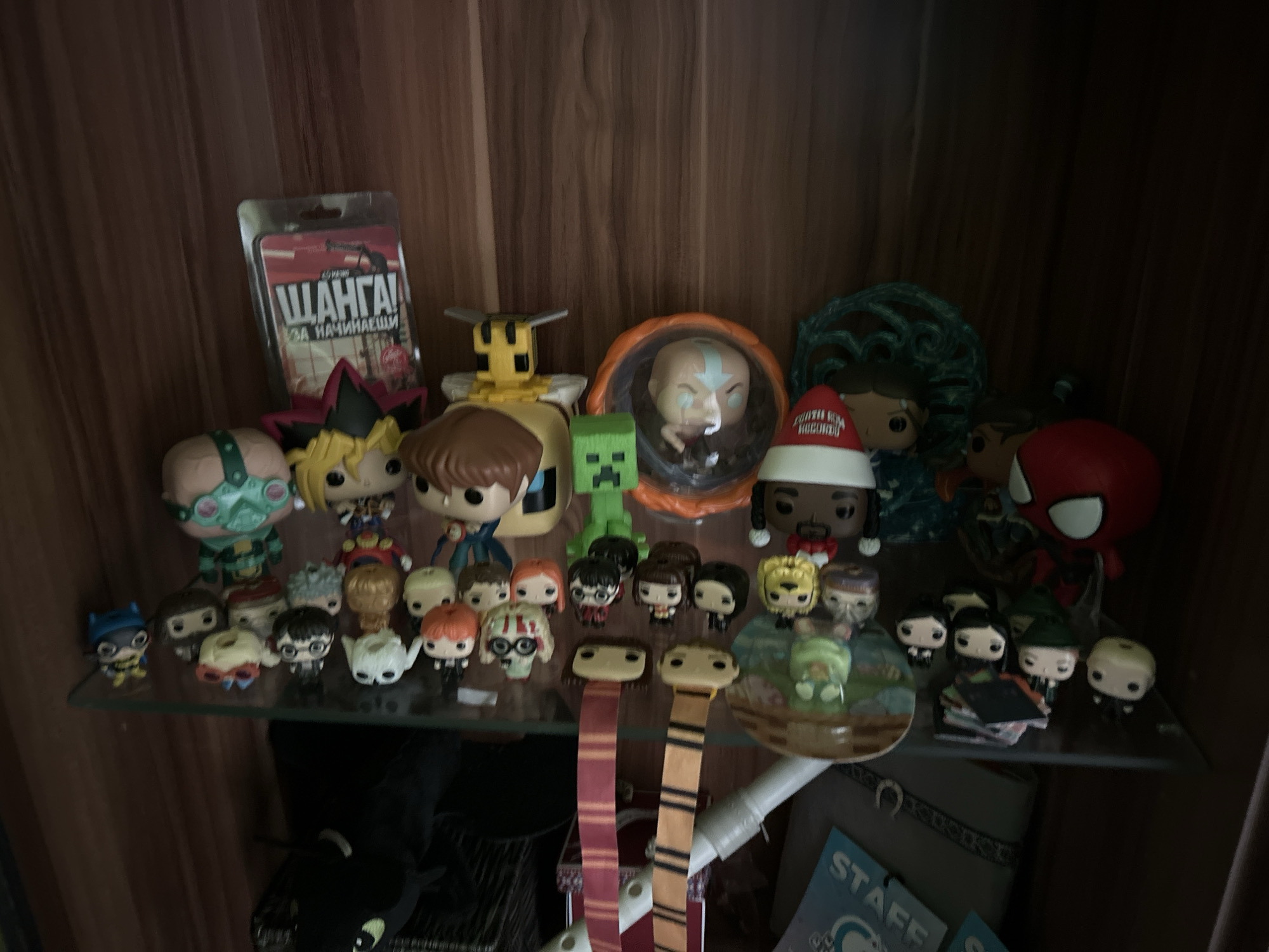 The image depicts a shelf filled with a variety of Funko Pop figures. The figures include characters from different franchises, such as Yu-Gi-Oh, Avatar: The Last Airbender, Minecraft, Harry Potter, and Marvel. There are also other objects on the shelf, such as a card game in Russian called "Shanga!", Hogwarts scarves, a plush toy, and books. The shelf is located inside a wooden cabinet. The location is not possible to tell from the visual information provided.