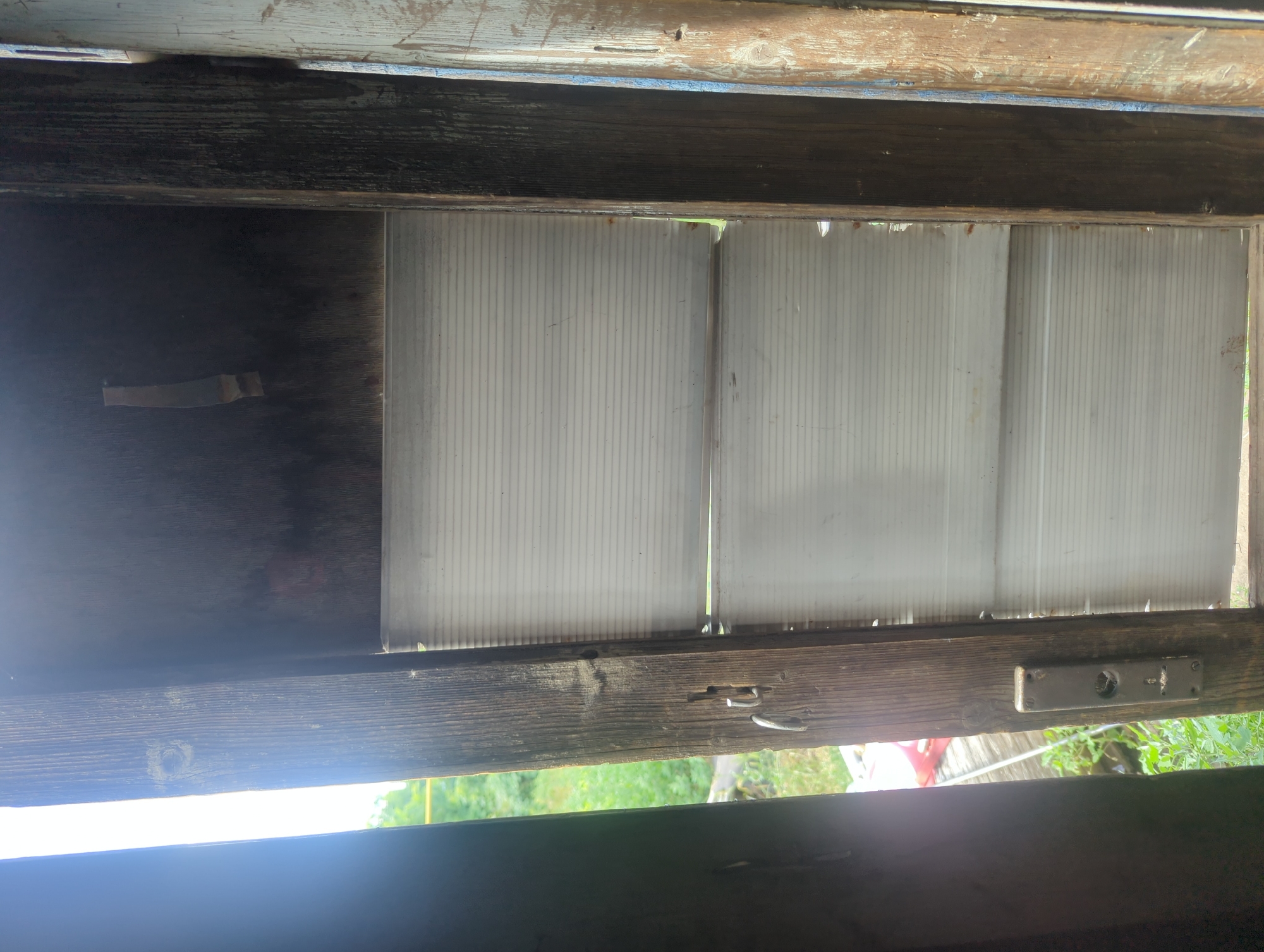 The image shows a view through an opening, possibly a window or door, in a weathered wooden structure. The opening is framed by rough, dark wooden beams. Through the opening, there are three panels of translucent white corrugated plastic, likely a makeshift window or roofing material. The plastic appears somewhat dirty and worn, with some damage visible along the bottom edge where it meets the lower wooden frame. To the left of the plastic panels, a dark wooden surface is visible, possibly a wall, with a piece of light-colored tape attached to it.
The lower part of the frame also shows wear and tear, with a metal latch or hook visible on the right side. Through the gaps at the bottom of the plastic panels and the opening below, glimpses of green foliage and trees can be seen, suggesting an outdoor setting. The lighting indicates it is daytime, with bright light coming from below and through the opening, creating some glare. The overall impression is of a rustic, perhaps abandoned or utilitarian structure. There is no visible text in the image.