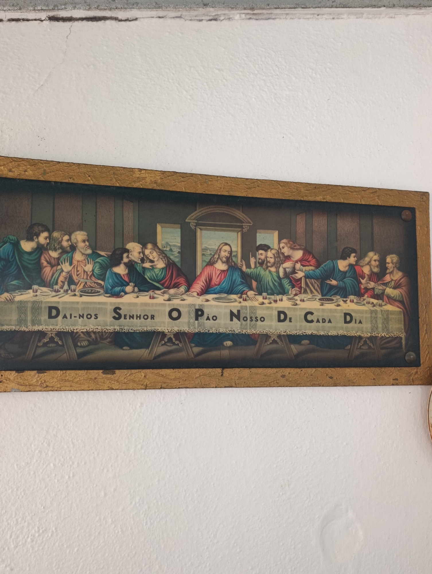 This is a framed artwork depicting the Last Supper, hanging on a white, textured wall. The painting shows Jesus at the center of a long table, surrounded by his twelve disciples. The disciples are seated on both sides of Jesus, engaged in conversation and eating. The table is covered with a white cloth, and there are plates, cups, and loaves of bread on it. The painting has Portuguese text written across the tablecloth that reads "DAI-NOS SENHOR O PAO NOSSO DE CADA DIA," which translates to "Give us this day our daily bread." The frame of the artwork is made of wood with a textured golden finish. The wall appears to be in an indoor setting, possibly a home or a place of worship. The lighting in the image is diffused, suggesting an interior light source. The artwork is positioned slightly off-center on the wall. Based on the style and religious iconography, this artwork is likely to be found in a Catholic household or church, potentially in a region with a strong Catholic tradition like Setúbal, Portugal.