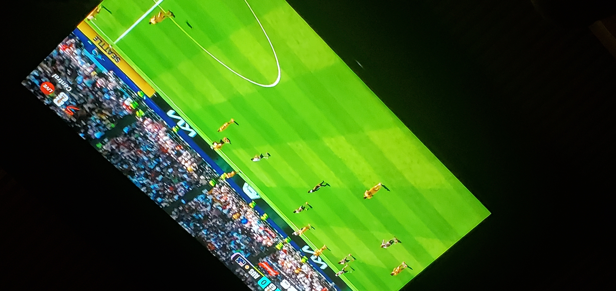 This is a photograph of a soccer match being broadcast on a television screen, angled from above. The scene displays a vibrant green soccer field with white markings. Several players in yellow and white uniforms are visible on the field. In the background, a stadium filled with spectators wearing a variety of colored clothing is depicted. Along the edge of the field, there is a yellow advertising board with the word "SEATTLE" in white lettering. A smaller advertising board with "KIA" is also visible. In the top corner of the screen, there is a broadcast graphic that includes the word "LIVE" in a red circle and "Central" in white text, along with a logo that appears to be "SABC". The broadcast also shows a score of "0" for "AUS". The image is captured in a dark environment, likely a room, with the television screen being the primary light source. There is no indication of the location being Gura Top, Nigeria.