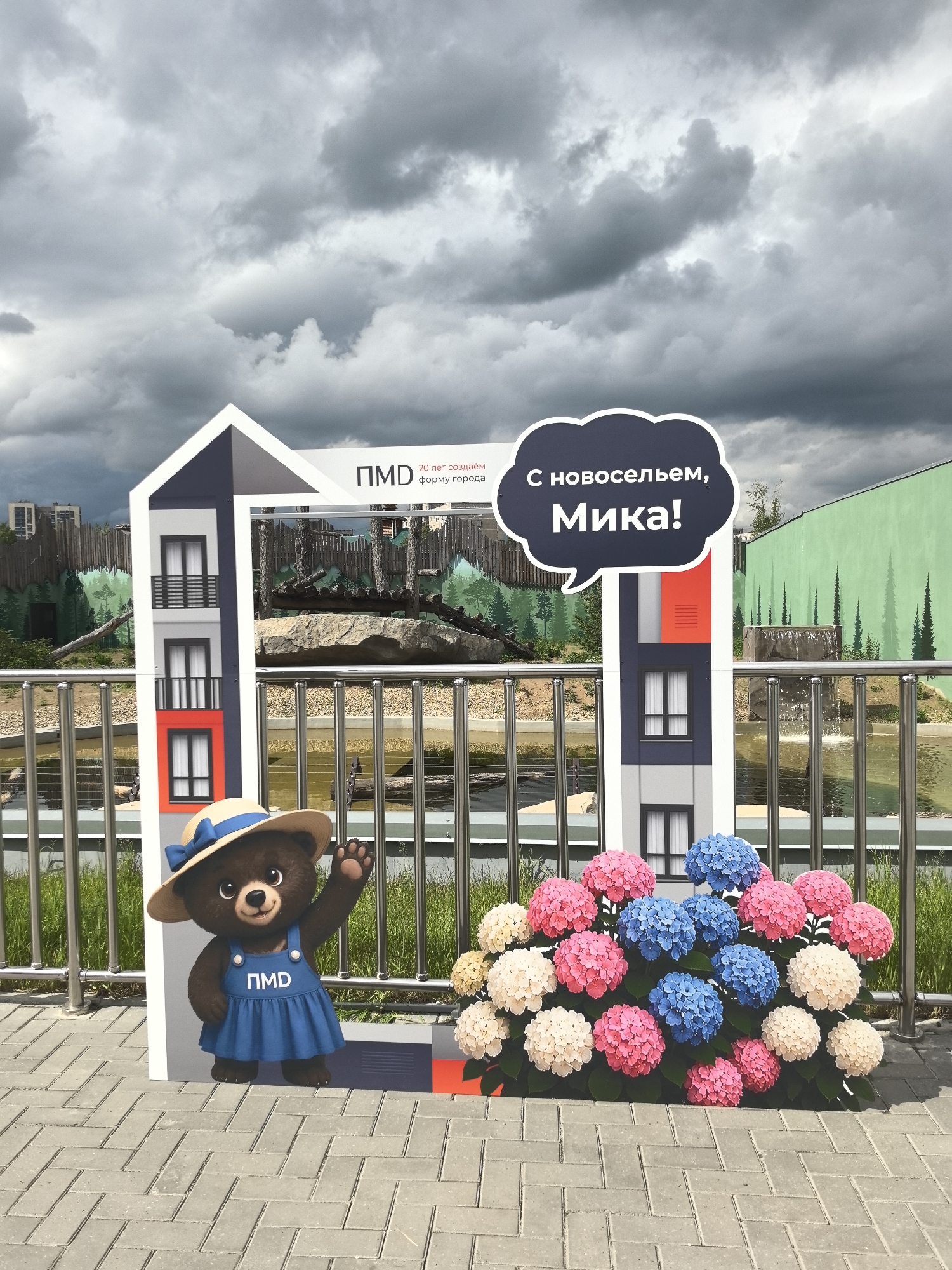 A photo shows a constructed house frame with a bear character in front of it. The character is a bear wearing a blue dress and a hat. A blue speech bubble to the right of the house structure reads, "С новосельем, Мика!". The backdrop consists of a green-painted wall with painted trees, a body of water behind a silver railing, and trees. The sky is cloudy. The house structure includes the letters "ПМД" above the slogan "20 лет создаём форму города." The house structure is decorated with pink, blue, and white hydrangeas at the bottom. This photograph was taken in the city of Perm, Russia.