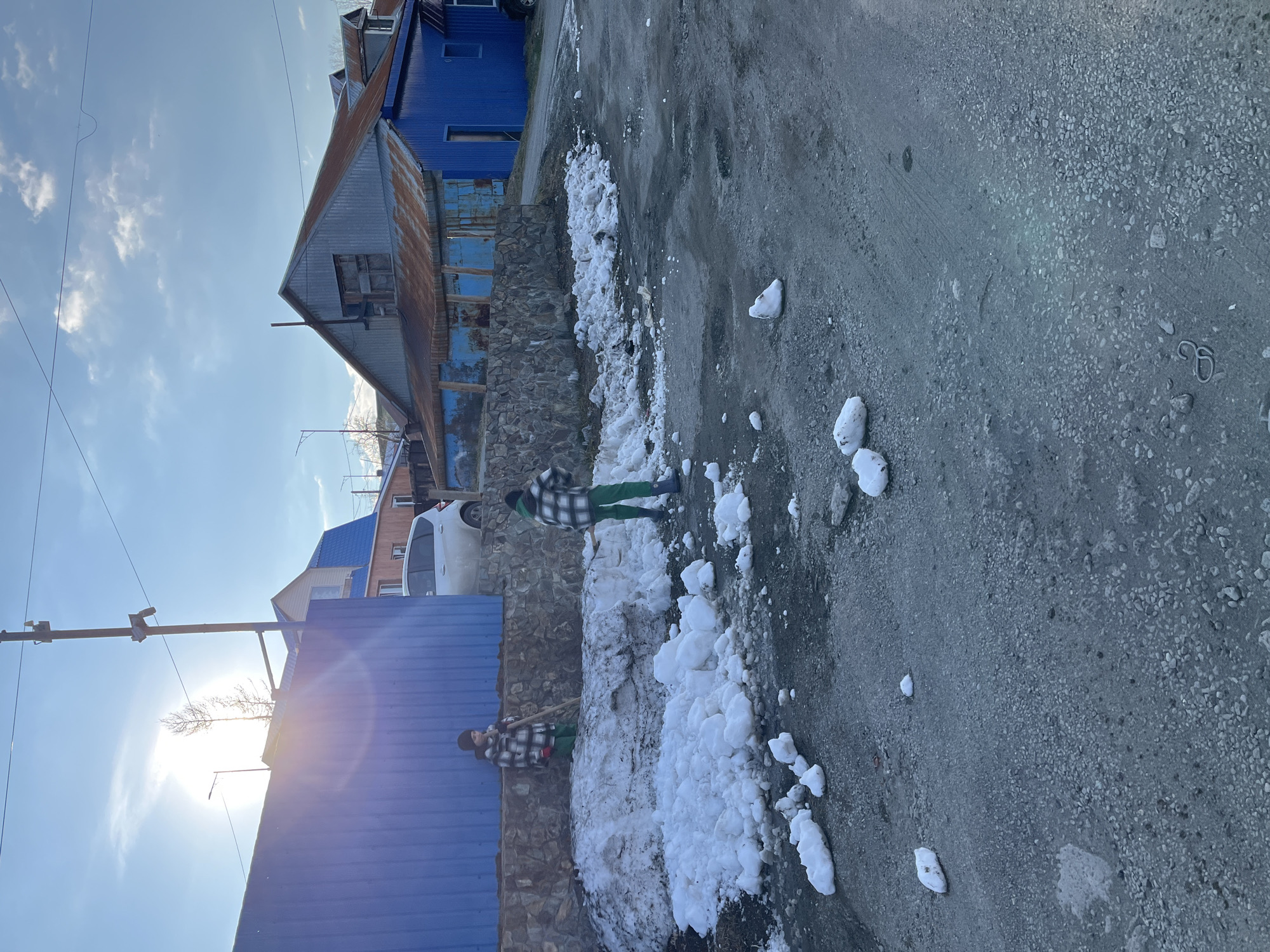 The image depicts a street scene in what appears to be a residential area. Two individuals are shoveling snow from a wall that separates the street from a property. The person closer to the camera is using a shovel to move snow from the wall towards the roadside. Snow has been partially cleared from the wall, and snow piles are visible on the ground. The street itself appears to be made of packed gravel. Buildings with blue painted surfaces are visible in the background along the left side of the image. A white car is also visible parked near one of the buildings. The sun is shining brightly and low in the sky. I cannot confirm that the location is the city of Petropavlovsk-Kamchatsky, Russia.