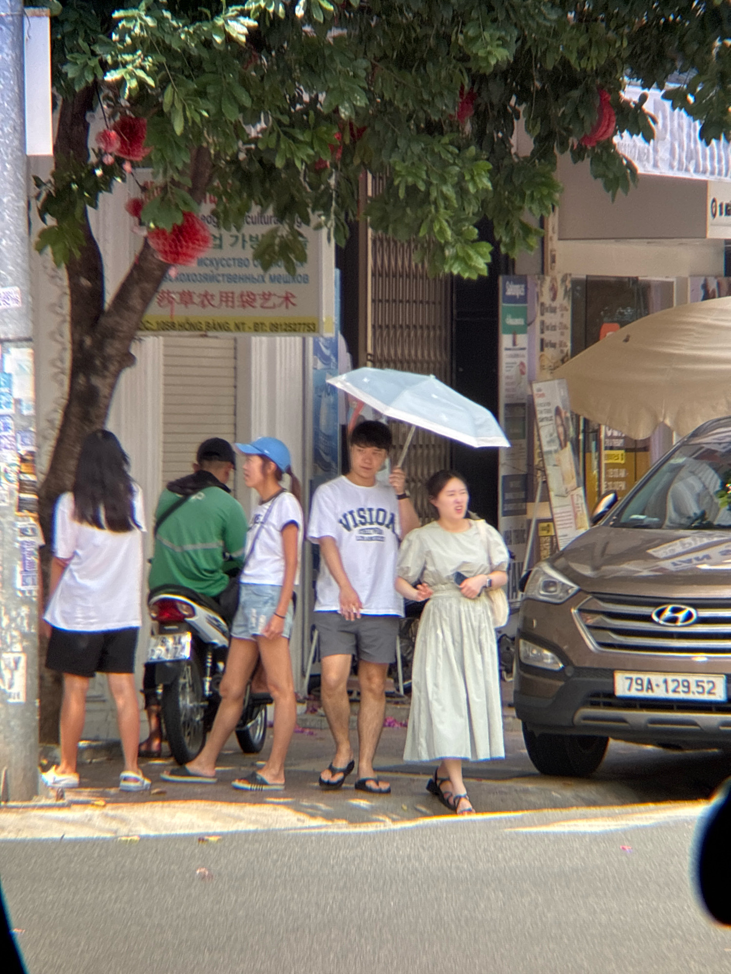 The image depicts a street scene in Nha Trang, Vietnam. In the foreground, a brown SUV is parked on the right side of the frame. Its license plate reads "79A-129.52". To the left of the SUV, a woman in a light-colored dress stands with an umbrella partially open. Next to her, a man wearing a "VISION" t-shirt and shorts stands under the umbrella. Further to the left, a group of three people stand near a motorcycle. One person, wearing a green jacket, is seated on the motorcycle. Two individuals, a woman in a white t-shirt and blue cap and another woman in a white t-shirt and black shorts, stand beside the motorcycle. A tree with red flowers is visible on the left side of the image, casting shadows on the pavement. The background shows buildings and shop fronts, consistent with a commercial street in a city.