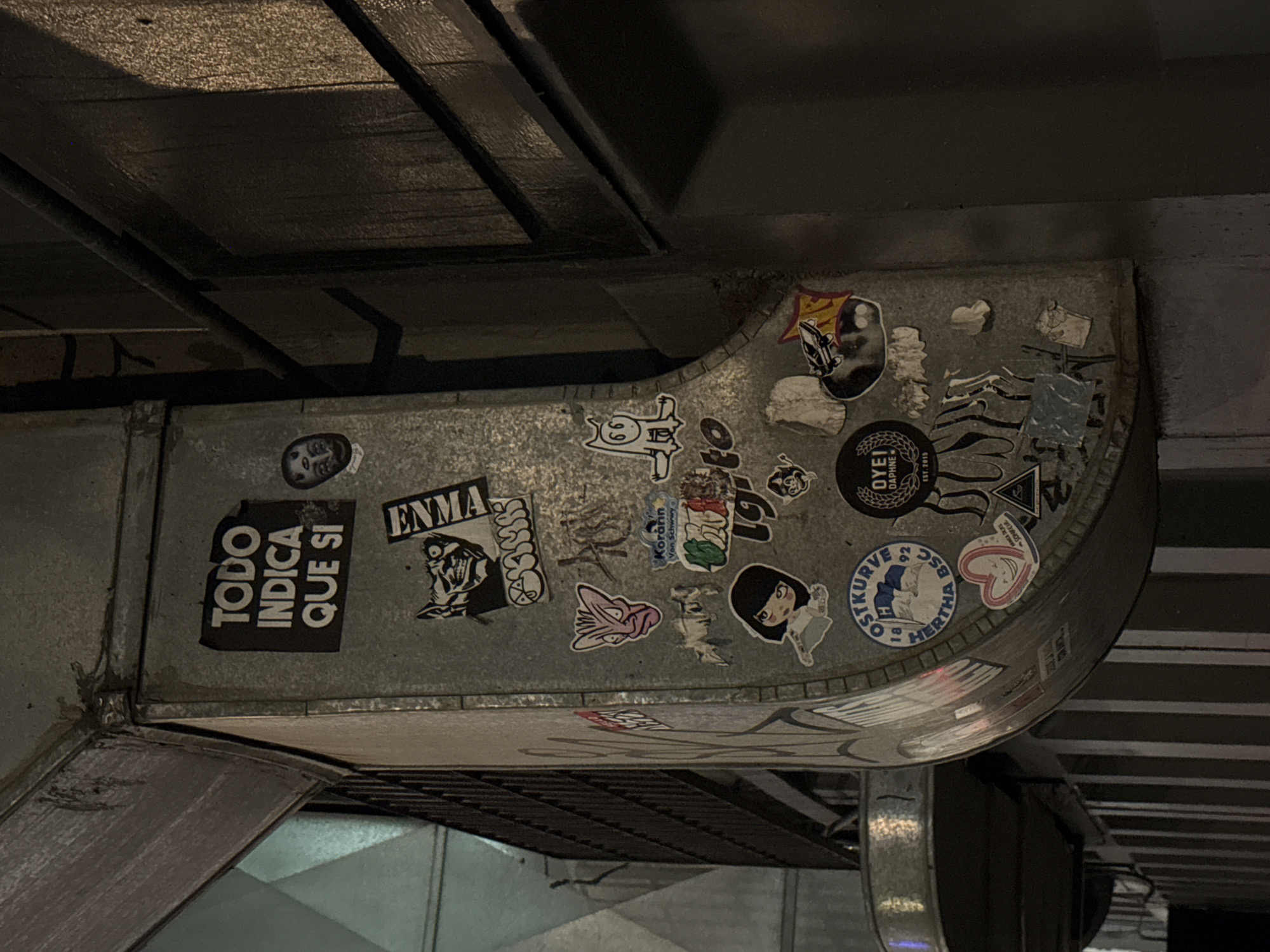 The image shows an indoor industrial setting, with a focus on a metal duct heavily covered in stickers. The duct is attached to the ceiling and likely part of the building's ventilation or air conditioning system. The stickers are of various designs, colors, and sizes, and appear to be a mix of logos, artwork, and text. The stickers include phrases like "TODO INDICA QUE SI", "ENMA", "STKURVE", "Korárin Von Schwwy", and "OYEI DAPHNE EST.2015". The environment is dark, with visible ceiling structures and the metallic surface of the duct reflecting light. The location in the city of Puebla, Mexico cannot be verified by the image alone.