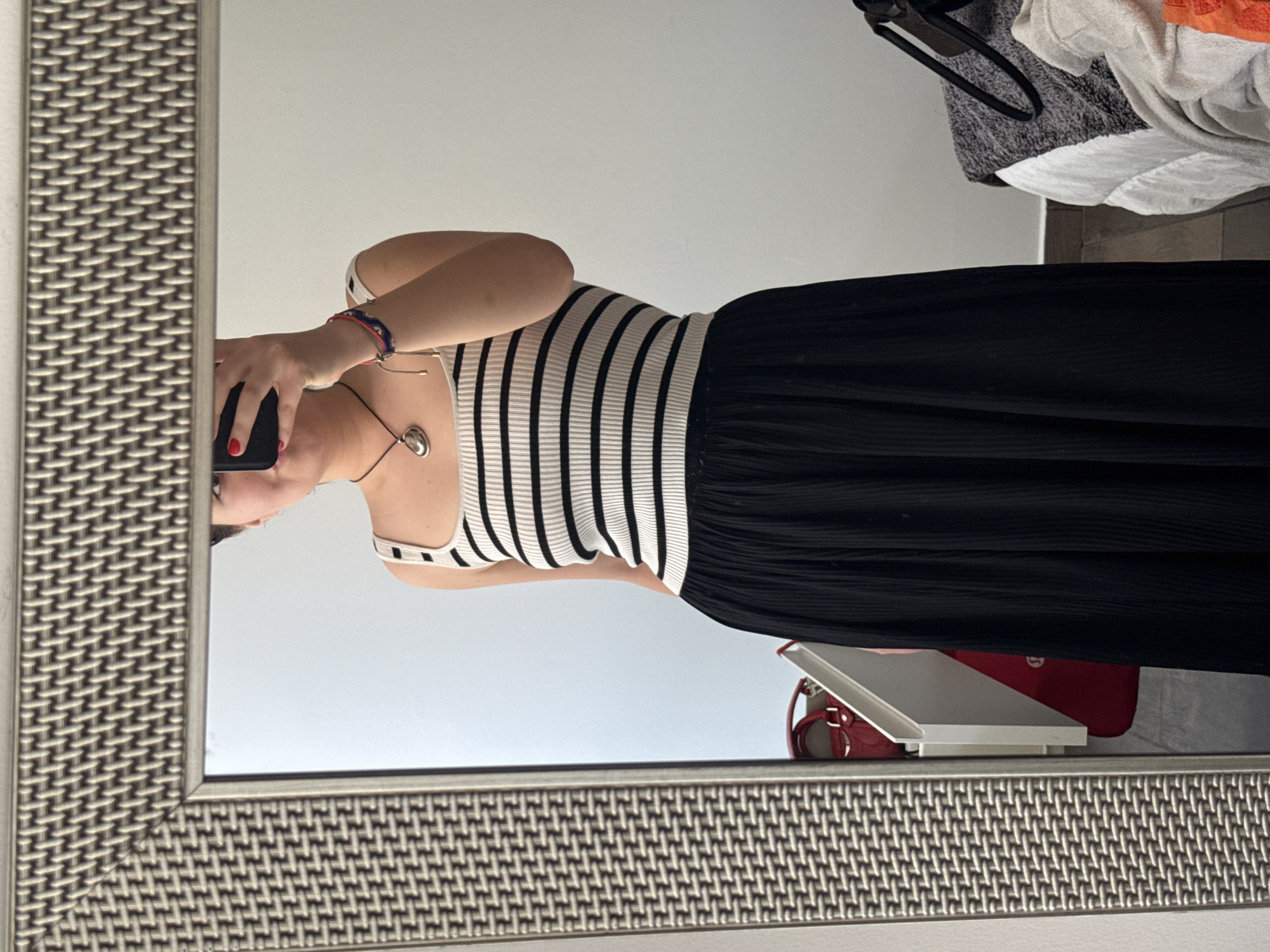 A medium shot captures a mirror selfie taken by a person, likely in an indoor setting in Mexico City, Mexico. The individual is positioned centrally, with their upper torso, arms, and part of their head visible in the mirror's reflection. They are wearing a sleeveless top with alternating white and black horizontal stripes, paired with a long, pleated black skirt.
The person's left hand, adorned with red nail polish, holds a dark-colored smartphone, indicating they are taking the picture. A multi-colored bracelet is visible on their left wrist, and a simple necklace with a silver-toned, rounded pendant hangs around their neck. The mirrored reflection shows a plain, light-colored wall as the background.
The mirror itself features a distinctive, textured frame with a woven or basket-weave pattern in a metallic or bronze hue, which is prominent along the left and bottom edges of the frame. Outside the mirror's reflection, to the upper right, a cluttered area shows folded textiles in shades of grey, white, and orange, along with a dark hanger. Below the reflection of the person's skirt, a bright red bag or purse is visible on the floor, next to a white, possibly plastic, stand or object. The flooring appears to be made of light-colored tiles or wood. The lighting suggests daytime. No discernible text is visible in the image.
