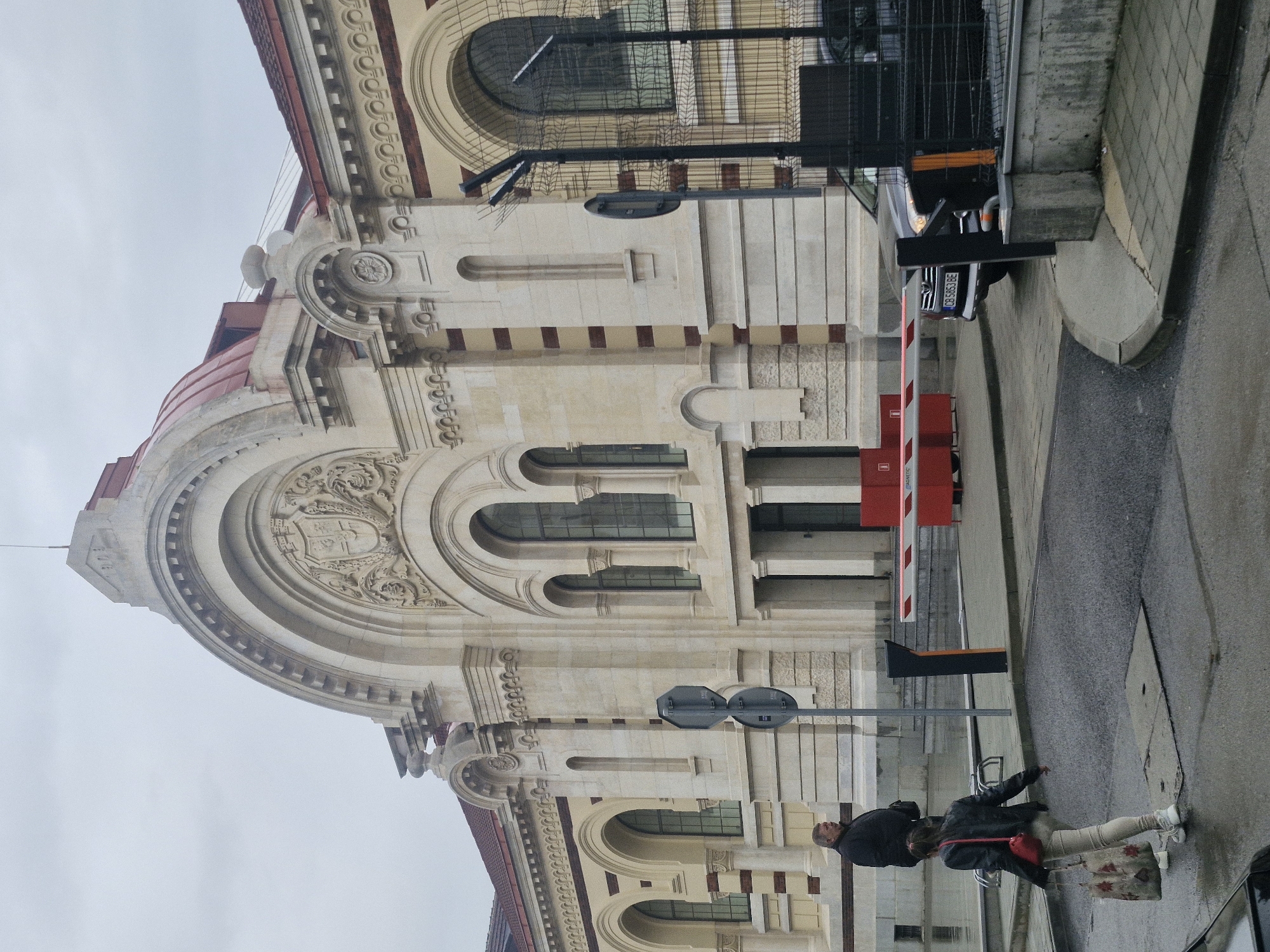 The image shows a partial view of the Central Sofia Market Hall located in Sofia, Bulgaria. The building's facade is prominently featured, showcasing its architectural details, including arches, decorative carvings, and red and cream color scheme.
A gray road runs along the right side of the building, where a couple is standing, and a parking barrier, painted white and red, sits just behind them. A dark-colored vehicle is partially visible beside the barrier. A street sign is visible between the couple and the building. The sky is overcast and gray.