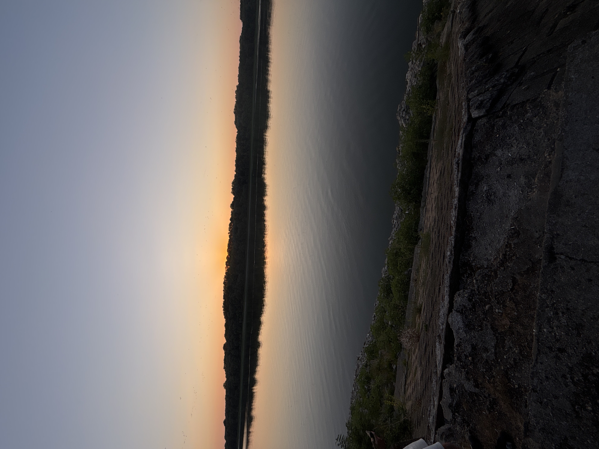 The image depicts a serene river scene at dusk. The sky transitions from a pale blue at the top to a warm orange and yellow hue near the horizon, suggesting the setting sun. A dense line of trees forms the far bank of the river, silhouetted against the colorful sky. The river itself is calm, with gentle ripples reflecting the light of the sky. The foreground shows a rocky embankment with some sparse vegetation. On the left side of the foreground, partially visible, is a person wearing white shorts. The image captures a moment of natural beauty and tranquility. There is no indication of the city of Marten, Bulgaria, in the image.