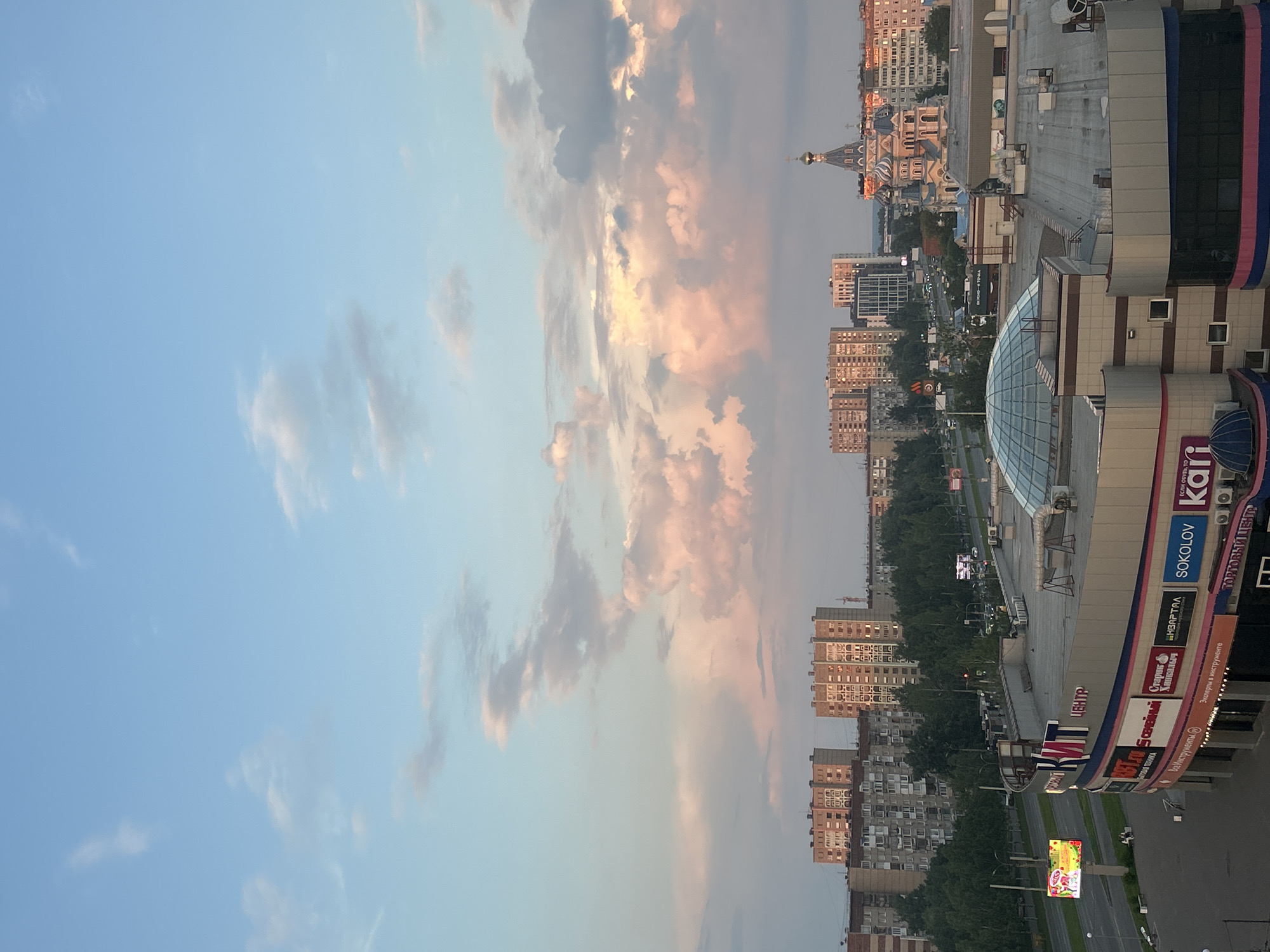An aerial, eye-level view captures a cityscape at dusk under a pale blue and pink sky filled with scattered clouds. In the foreground, a multi-story commercial building with a rounded section topped by a glass dome dominates the lower right. The building features signage with Cyrillic lettering, including "ЦЕНТР" and "SOKOLOV," along with various other brand names. To the left of this building, a busy road with vehicles is visible, bordered by a sidewalk with a few pedestrians. A large electronic billboard displaying colorful advertisements is situated to the far left.
Further into the background, a dense line of trees separates the foreground structures from a row of apartment buildings, characterized by their uniform architecture and multiple windows. Behind the apartment buildings, a prominent Orthodox church stands out with its ornate design, featuring multiple domes, the largest of which is gilded. The sky above transitions from a clear blue near the top to hues of pink and orange where it meets the horizon, illuminated by the setting sun. The overall scene depicts a blend of residential, commercial, and religious architecture within an urban setting.