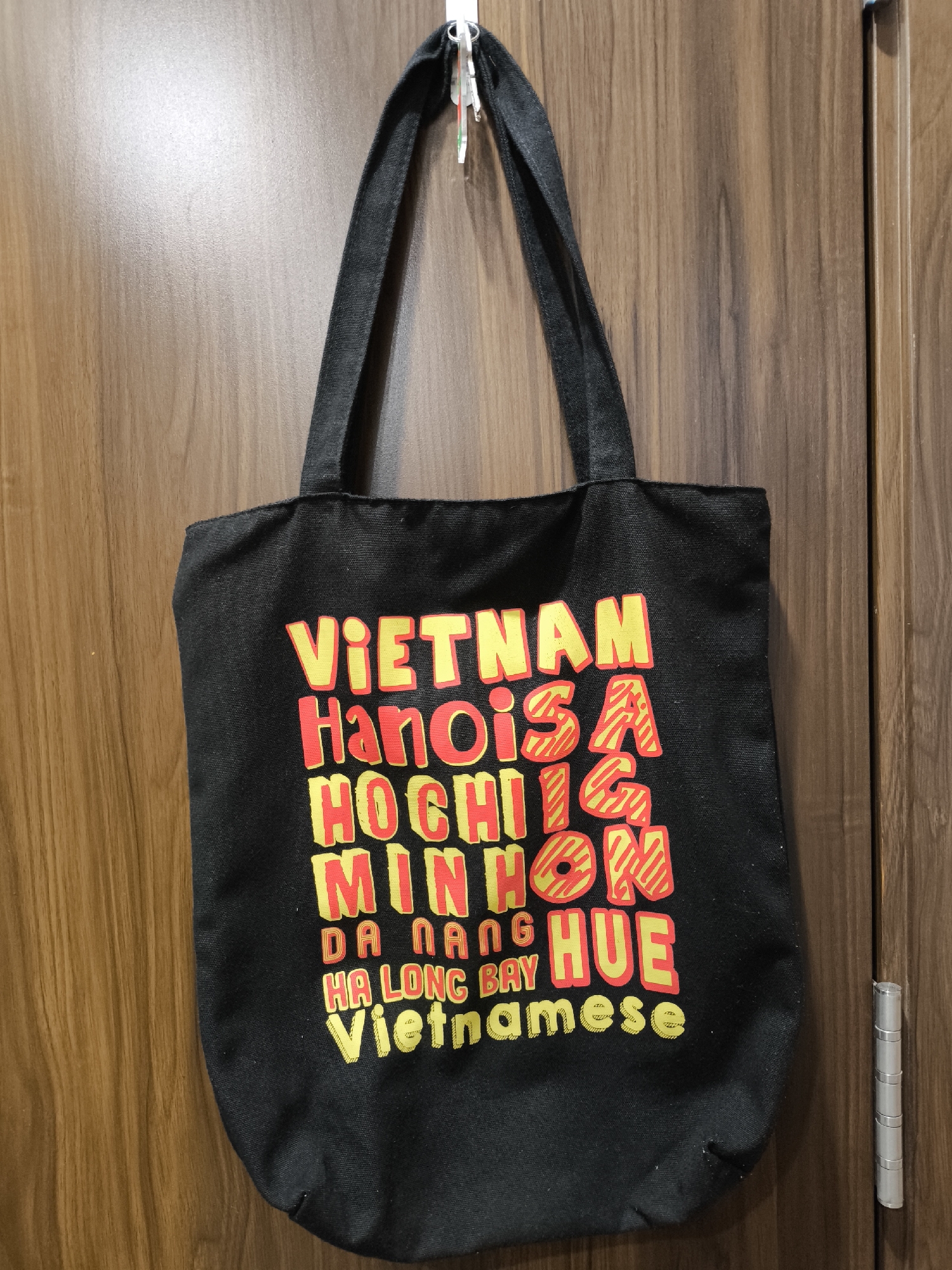 A black canvas tote bag with red and yellow lettering is hanging on a wooden door. The lettering on the bag reads: "VIETNAM HanoiSAGOCHIH MINHOM DANANG HUE HALONG BAY Vietnamese." The bag is suspended by a white hook, and a silver door hinge is visible on the right side of the image. The environment is indoors, with the wooden door filling the background. The city of Đà Nẵng is mentioned on the bag as a point of interest in Vietnam.