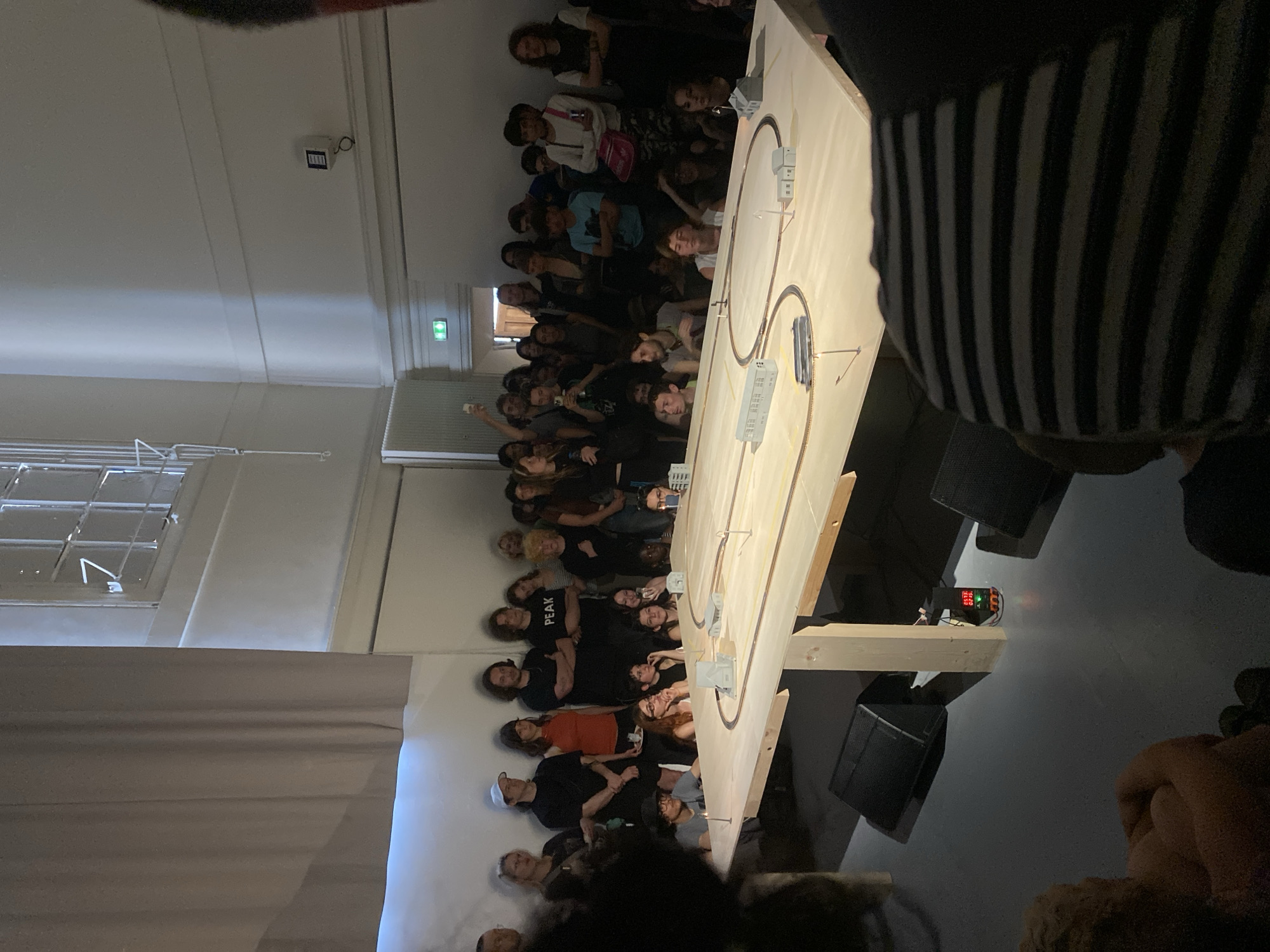 An indoor scene depicts a large group of people gathered in what appears to be an exhibition or installation setting. A wooden structure occupies the center of the space, featuring a model train track with small buildings placed alongside it. A digital display on the structure shows numerical data. People are standing and looking toward the structure. The walls are white, with visible windows, air conditioning and emergency lighting. The overall lighting in the room is moderate. The location appears to be in Paris, France.