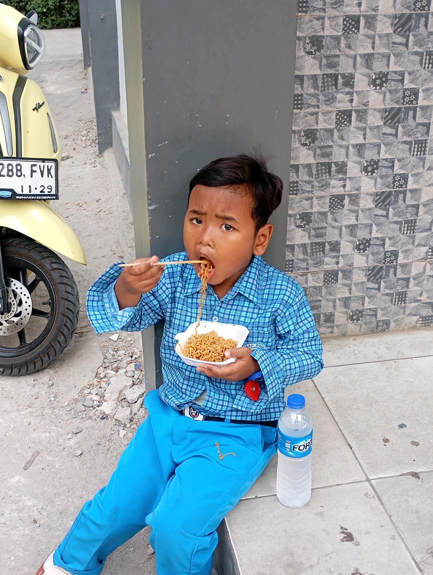 A young boy in a blue plaid shirt and bright blue pants sits on a concrete ground, eating noodles with chopsticks from a white styrofoam container. A bottle of Fort-W water is to his right, and a yellow scooter is parked to his left. The background consists of a gray wall with patterned tiles and a grey pillar. The scene appears to be outdoors, possibly in front of a building, in the city of Babelan, Indonesia.