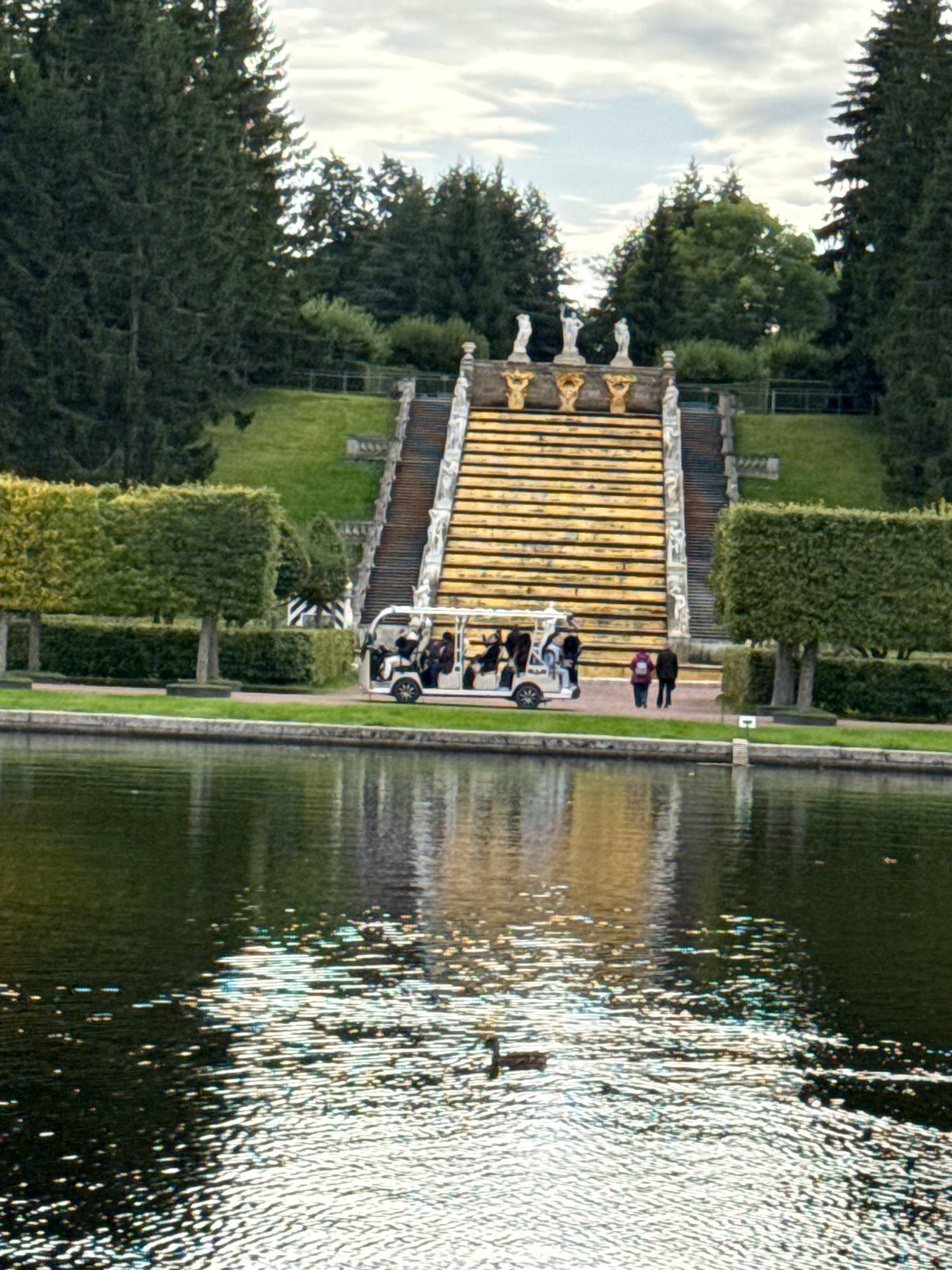 The image depicts a sunny day scene at the Grand Cascade in Peterhof, Russia, a UNESCO World Heritage site. In the foreground, a calm body of water reflects the surrounding scenery, with sunlight creating sparkling patterns on its surface. A duck is visible swimming in the water.
Dominating the middle ground is a large, ornate golden staircase, characteristic of Peterhof's fountains and gardens. At the base of the staircase, a white electric cart, resembling a golf cart, carries several passengers, suggesting a tourist attraction. Two individuals, a woman with a red backpack and a man in a dark jacket, stand near the cart.
The background features meticulously manicured green hedges and trees, indicative of formal gardens. Further up, more of the grand staircase is visible, adorned with statues and architectural elements. Towering evergreen trees flank the scene, with a cloudy sky overhead. The overall impression is one of grandeur and natural beauty, highlighting a popular tourist destination.