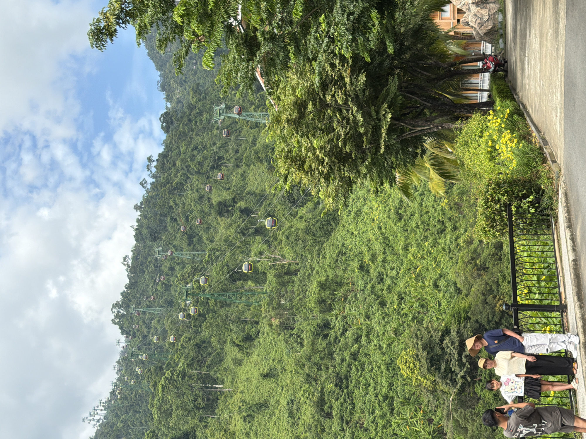 This is a photograph of a cable car system ascending a densely forested mountainside in the city of Đà Nẵng, Vietnam. The cable cars, which are yellow and red, are suspended from green pylons and traverse the steep, lush green terrain. In the foreground, a group of four people are standing near a black metal fence. A man on the left is holding a phone, appearing to take a photo. Next to him are two women and a man wearing a straw hat. They are standing on a paved walkway with yellow flowers and green bushes bordering it. Further in the background, there is a building with orange accents and a rock formation. The sky is partly cloudy with patches of blue.