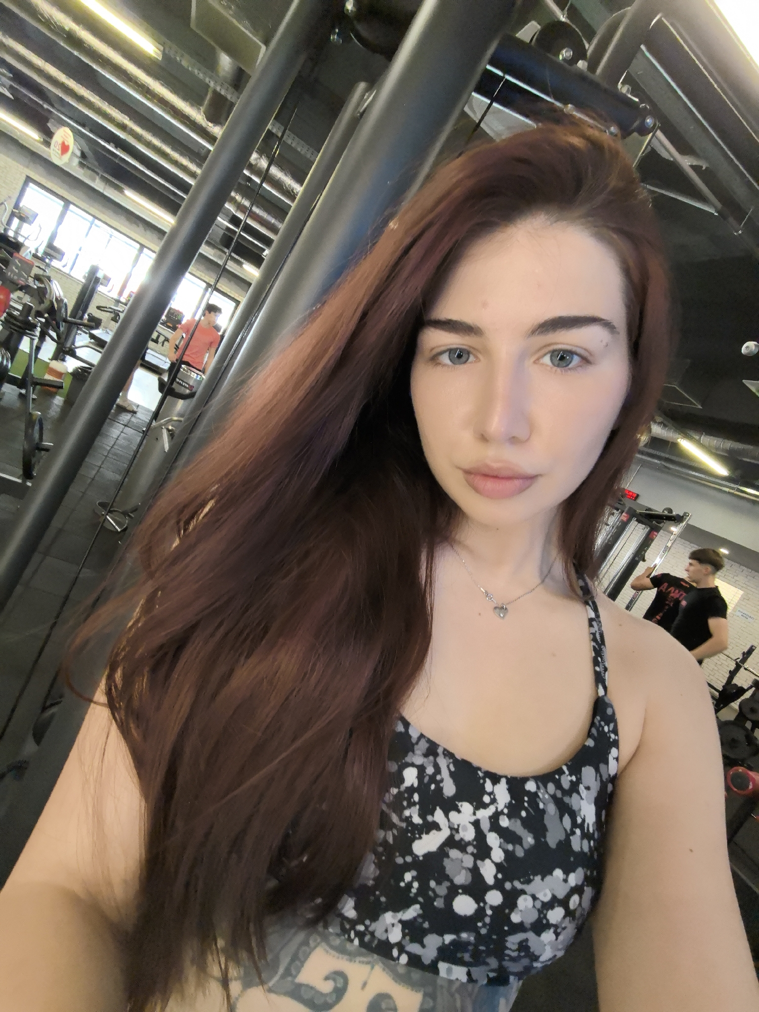 A woman with long red hair, wearing a black and white patterned sports bra and a silver heart necklace, poses for a selfie in a gym. The gym appears to be well-lit and contains various exercise machines and weights. Another person in a black shirt is visible in the background. The location is possibly in the city of Stara Zagora, Bulgaria.