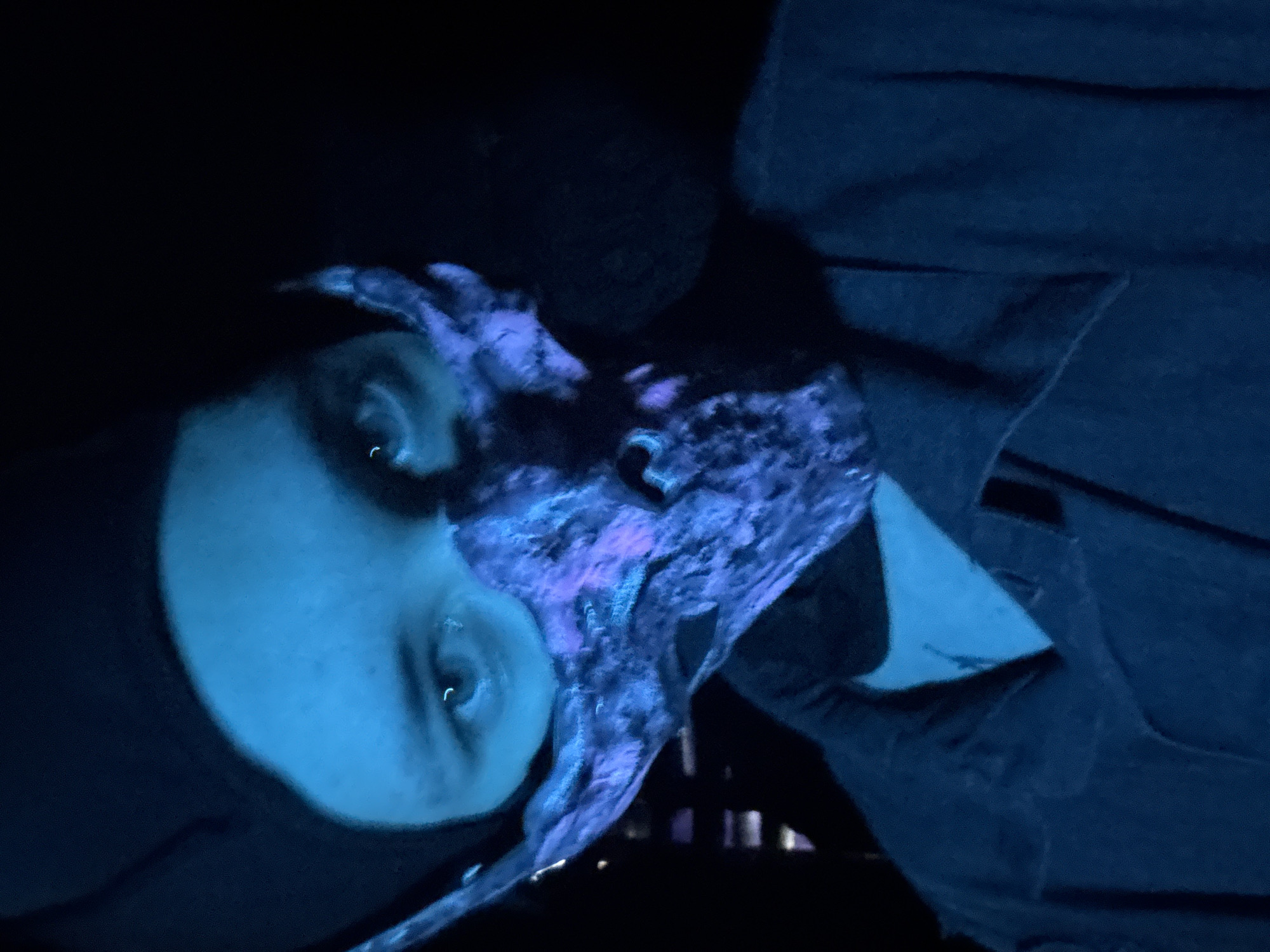 A person's face, illuminated in a blue light with a slight purple hue on the right side, is visible. The person has a dark shirt, and the right side of the frame shows a dark blue jacket. In the background, some faint vertical lines suggest a fence or similar structure. The image does not show any elements that could relate to the city of Saratov, Russia.