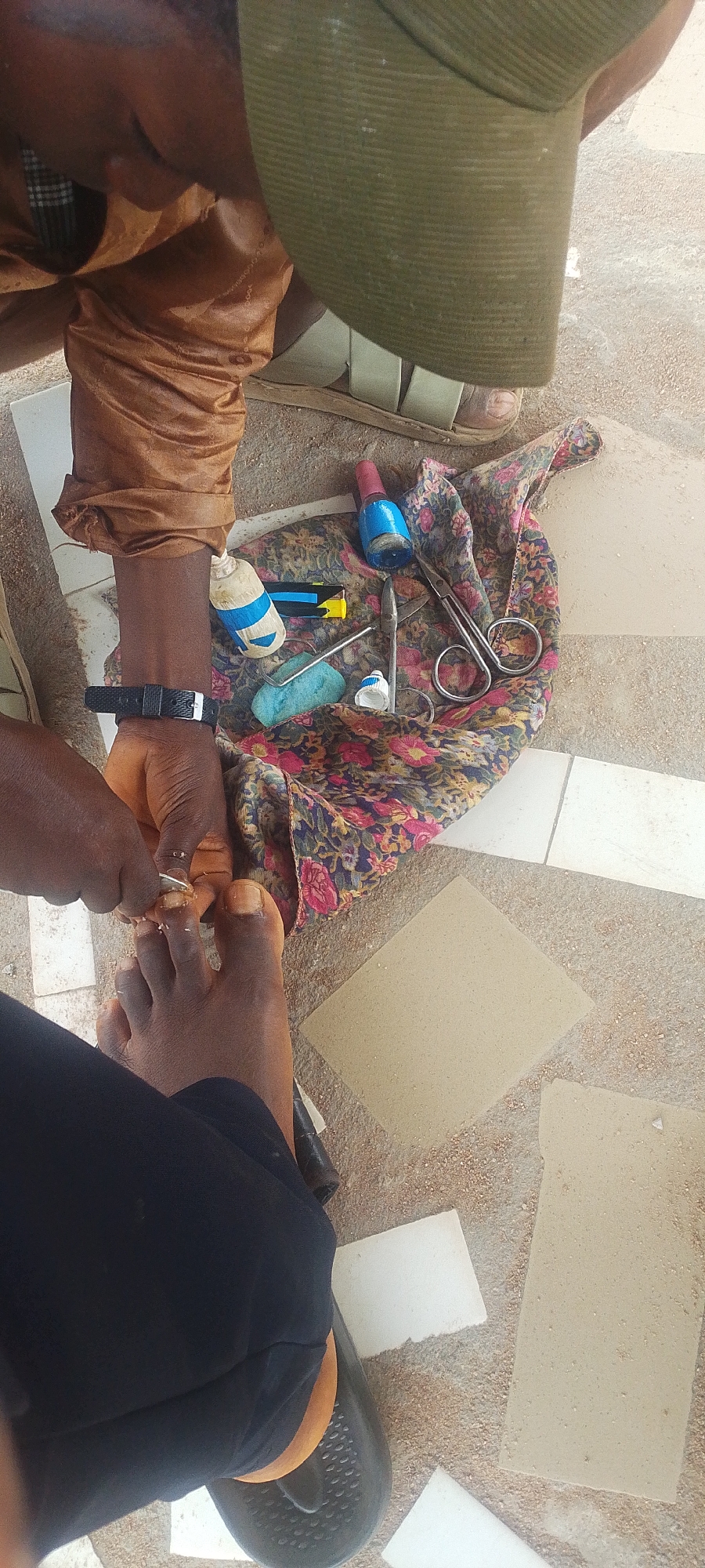 An outdoor scene in Jalingo, Nigeria, shows a person's feet and hands engaged in a pedicure. A person, likely female based on the nail polish and the context of the activity, is seated and performing the pedicure on another person's feet. The person receiving the pedicure is on the ground, wearing dark pants and a black sandal. The pedicurist is wearing a brown shirt and a green cap. Tools for the pedicure, including nail clippers, scissors, nail polish, and a buffer, are laid out on a floral cloth on the ground. The ground is a mixture of sand and broken tiles. The lighting suggests it is daytime.