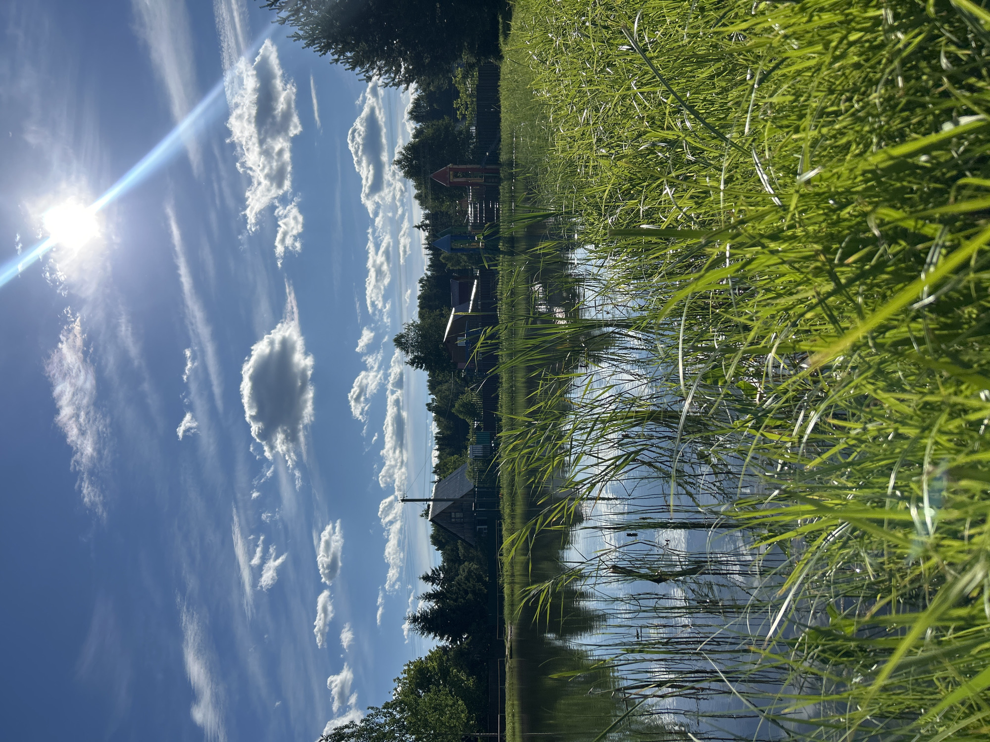 In the city of Слава Снт, Russia, a body of water is framed by tall, green grass in the foreground, with reflections of the sky and surrounding trees visible on its surface. Across the water, a dense line of trees serves as a backdrop for several structures, including a house with a dark roof and a visible chimney, and a colorful playground with red and blue rocket-shaped structures. The sky above is a vibrant blue, dotted with fluffy white clouds and illuminated by the bright sun, which casts a strong lens flare. The overall scene depicts a peaceful, rural setting during daylight.