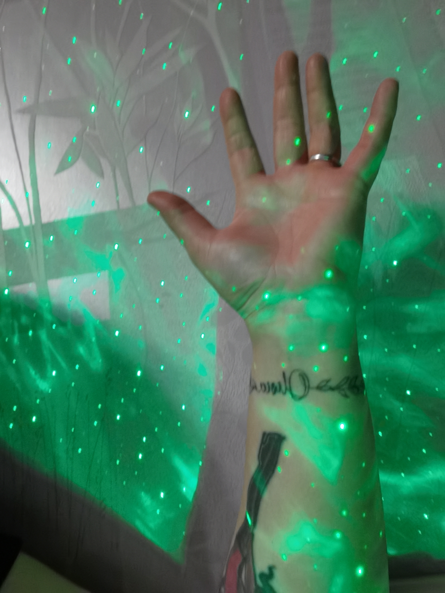 A close-up shot of a person's arm and hand in front of a fabric with a floral pattern, illuminated by green laser lights. The arm has two visible tattoos, one on the wrist and one further up the forearm. The hand is open, with a ring visible on one finger. The location in Votkinsk, Russia is unknown.