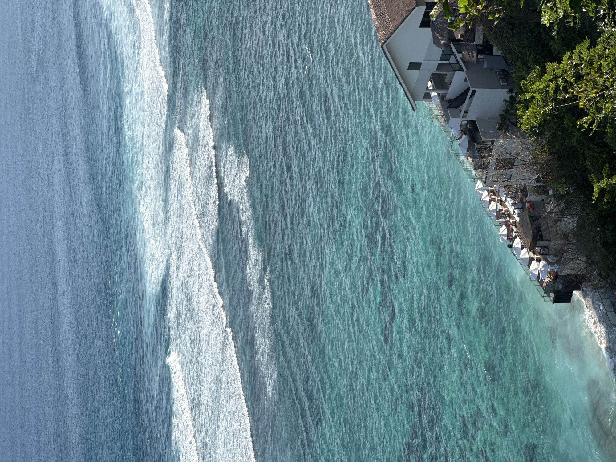 An aerial, eye-level view of the ocean with a coastal resort is shown. The ocean is a bright turquoise color with white-capped waves breaking. The resort is built on the side of a cliff overlooking the ocean. It has several levels with white buildings, umbrellas, and seating areas. There are some people on the balconies and at tables. Lush green trees are visible in the foreground. The image appears to be taken on a sunny day. This scene is located in Kuta Selatan, Indonesia.