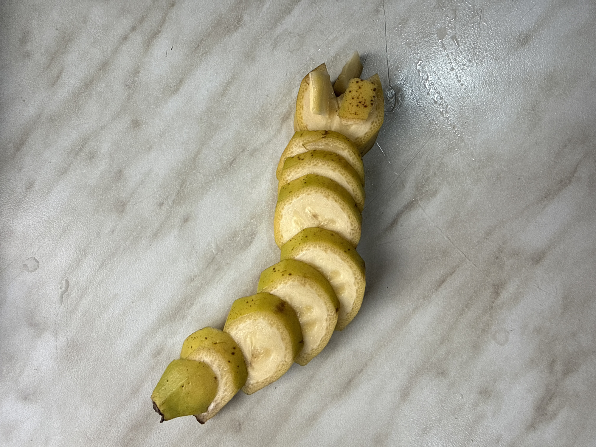 A slightly browned banana, cut into several slices, lies on a speckled, gray-and-white countertop. The banana is arranged in a curved line, with the top end pointing left and the bottom end pointing right. The slices are approximately half an inch thick and are arranged overlapping each other. The countertop appears to be wet in some areas, with small droplets of liquid visible.