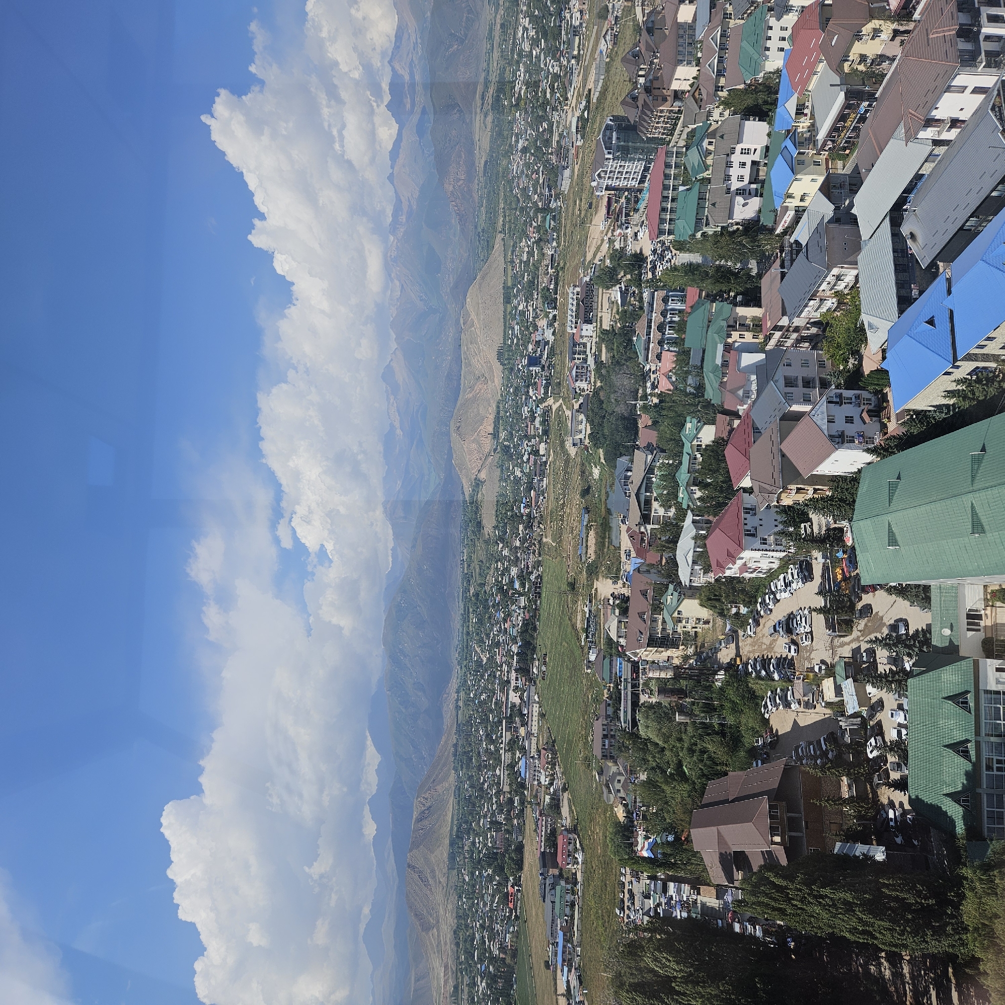The image is an aerial view of the city of Bosteri, Kyrgyzstan, on a clear day with a blue sky and scattered white clouds. The city is nestled in a valley with rolling, arid hills and a majestic mountain range in the background. The foreground and midground are densely populated with a variety of buildings, including residential homes and larger structures, many with colorful roofs. Green trees and vegetation are interspersed throughout the urban landscape. Roads are visible, lined with parked cars. The overall scene depicts a thriving, albeit compact, settlement against a dramatic natural backdrop.