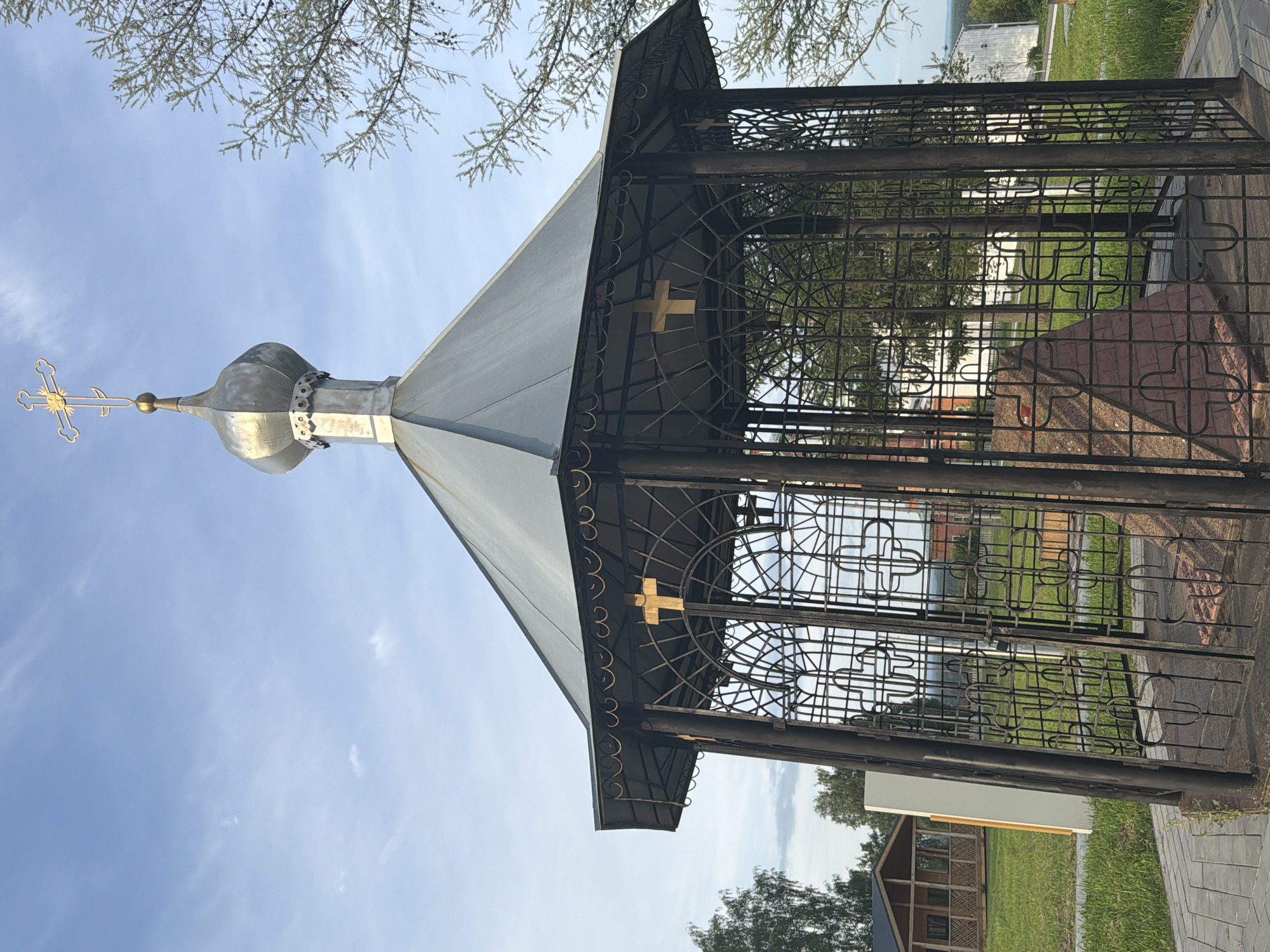 The image depicts a small, octagonal gazebo with a domed roof and a golden cross at its apex, located in Cherdyn, Russia. The gazebo is constructed of dark metal latticework, enclosing a stone monument or altar. The monument is pyramid-shaped and made of reddish-brown material, possibly brick or stone. There are golden crosses visible on the exterior of the gazebo's railings. The surrounding environment includes a paved pathway leading up to the gazebo, a well-maintained grassy area, and trees. In the background, a small wooden building with a porch is visible. The sky is clear with a few wispy clouds. The scene appears to be a religious site or a memorial.