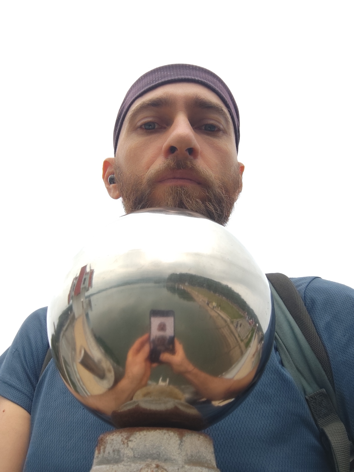 A selfie, taken from a low angle, shows a man with a beard and a dark purple headband, holding a reflective sphere. His blue shirt and backpack are visible. Reflected in the sphere is a water body, a walkway, and a building with a red roof. A person is also reflected holding a mobile phone. The sphere sits on a weathered, stone-like structure. The image was taken at the Xuan Huong Lake in Da Lat, Vietnam.
