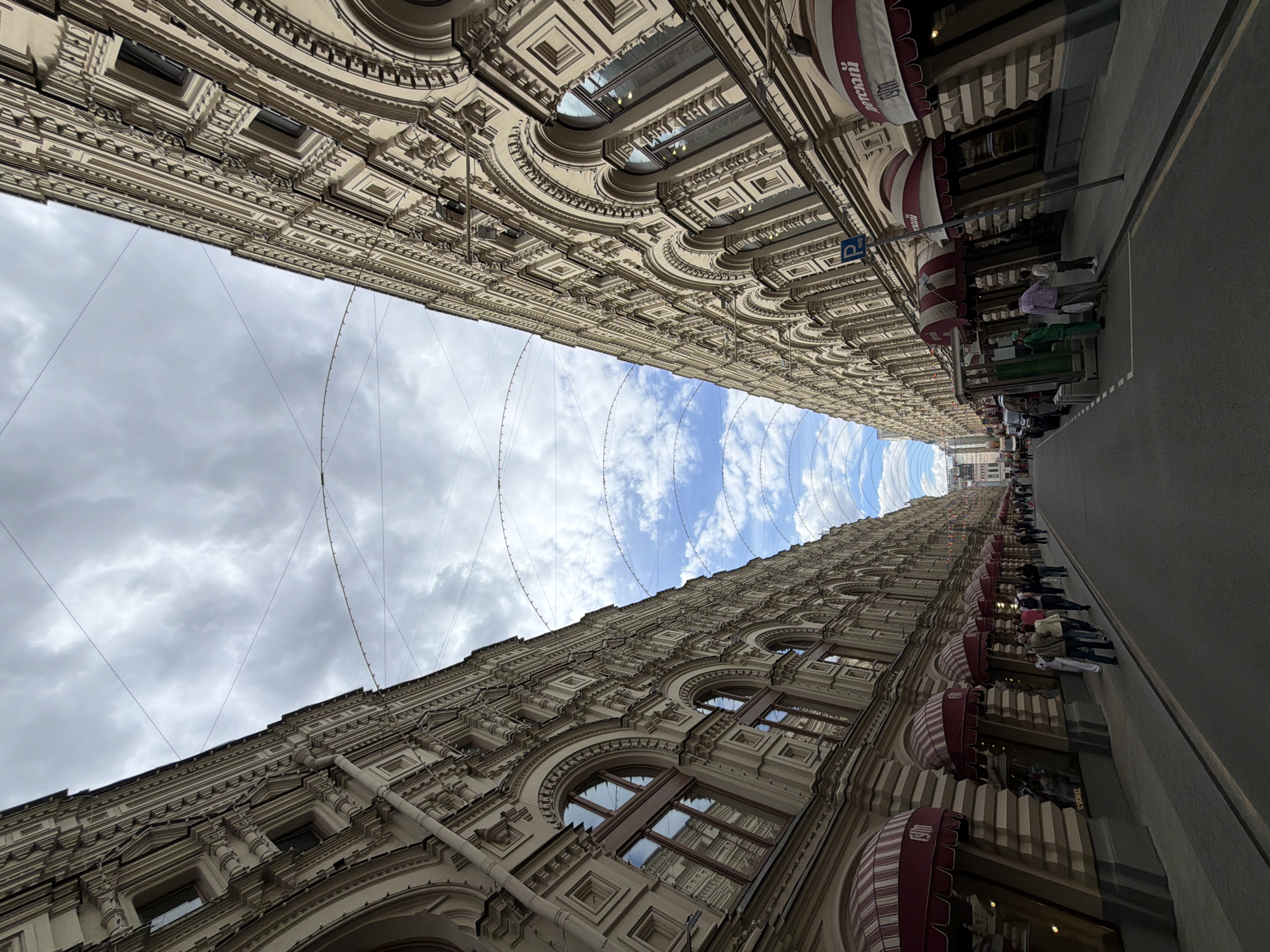 The image depicts a low-angle, perspective view of two ornate buildings facing each other across a narrow street in Moscow, Russia. The sky is visible between the buildings, featuring a mix of blue sky and scattered clouds. The buildings are tall and have elaborate architectural details, including arched windows, decorative moldings, and awnings over shopfronts. People are seen walking along the sidewalk next to one of the buildings. The overall impression is of a bustling urban environment with a rich history and architectural grandeur.